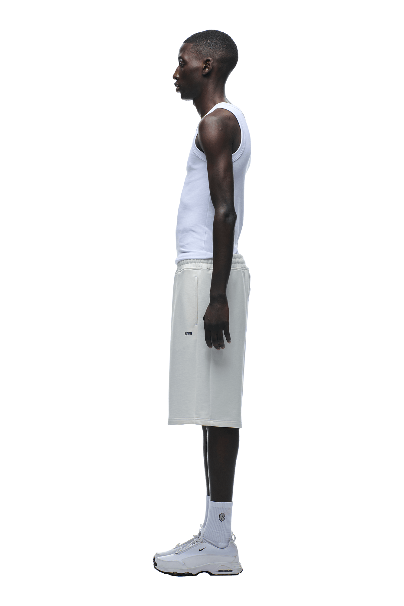 A person in profile faces left, wearing 6PM+ SHORTS COCONUT WHITE by 6PM with a relaxed white tank top, white socks, and sneakers, set against a plain green background.