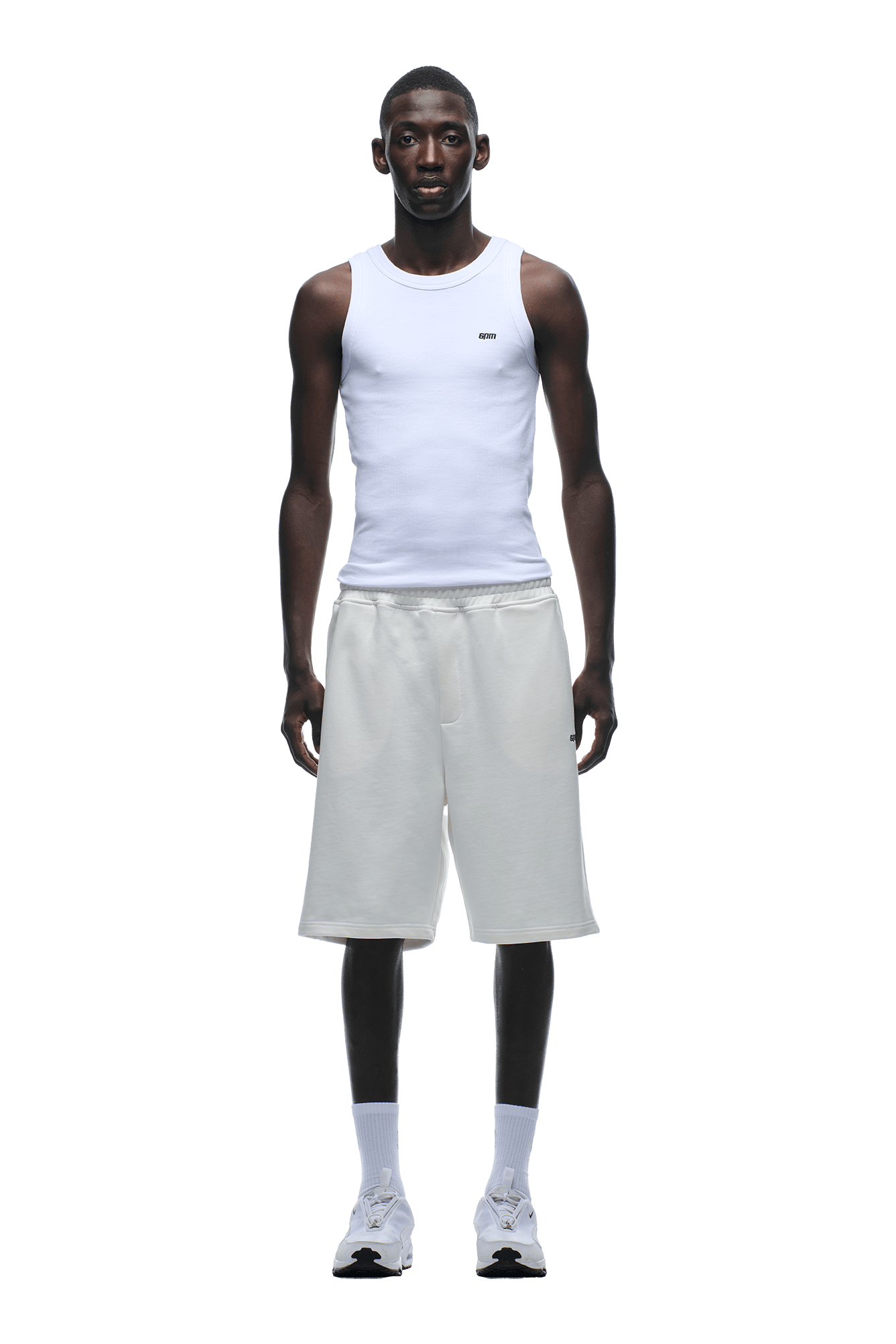 A man stands facing forward, wearing a relaxed-fit white sleeveless tank and 6PM+ SHORTS COCONUT WHITE by 6PM with a drawstring waist, paired with white socks and sneakers, against a plain background.