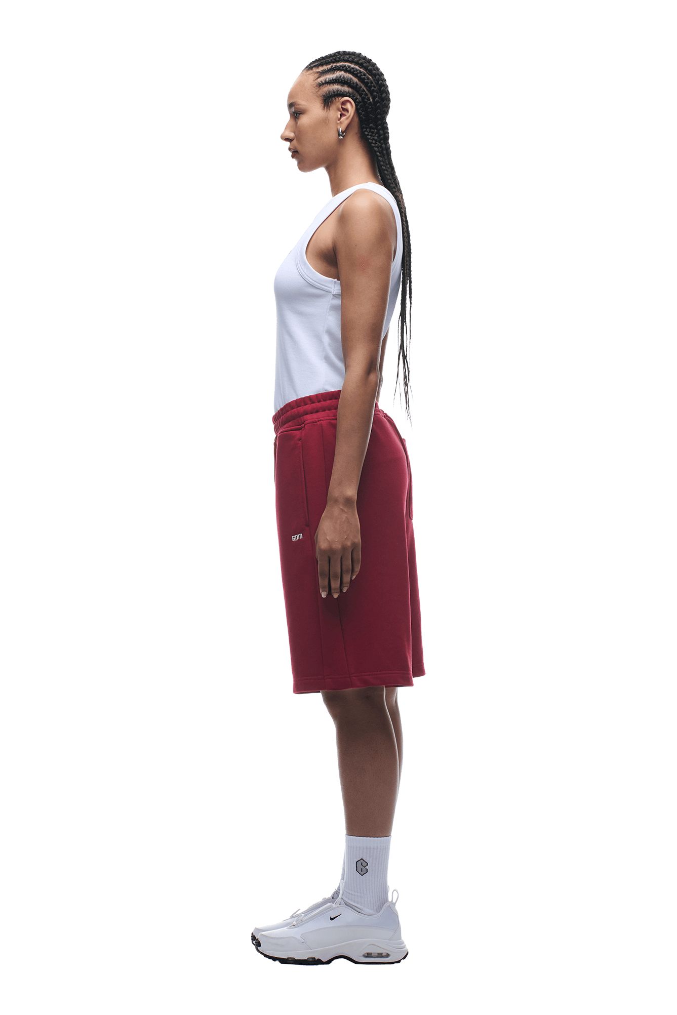 A woman with long braided hair stands in profile against a plain background, wearing a white tank top and 6PM 6PM+ SHORTS WINE RED, paired with white crew socks and white sneakers.