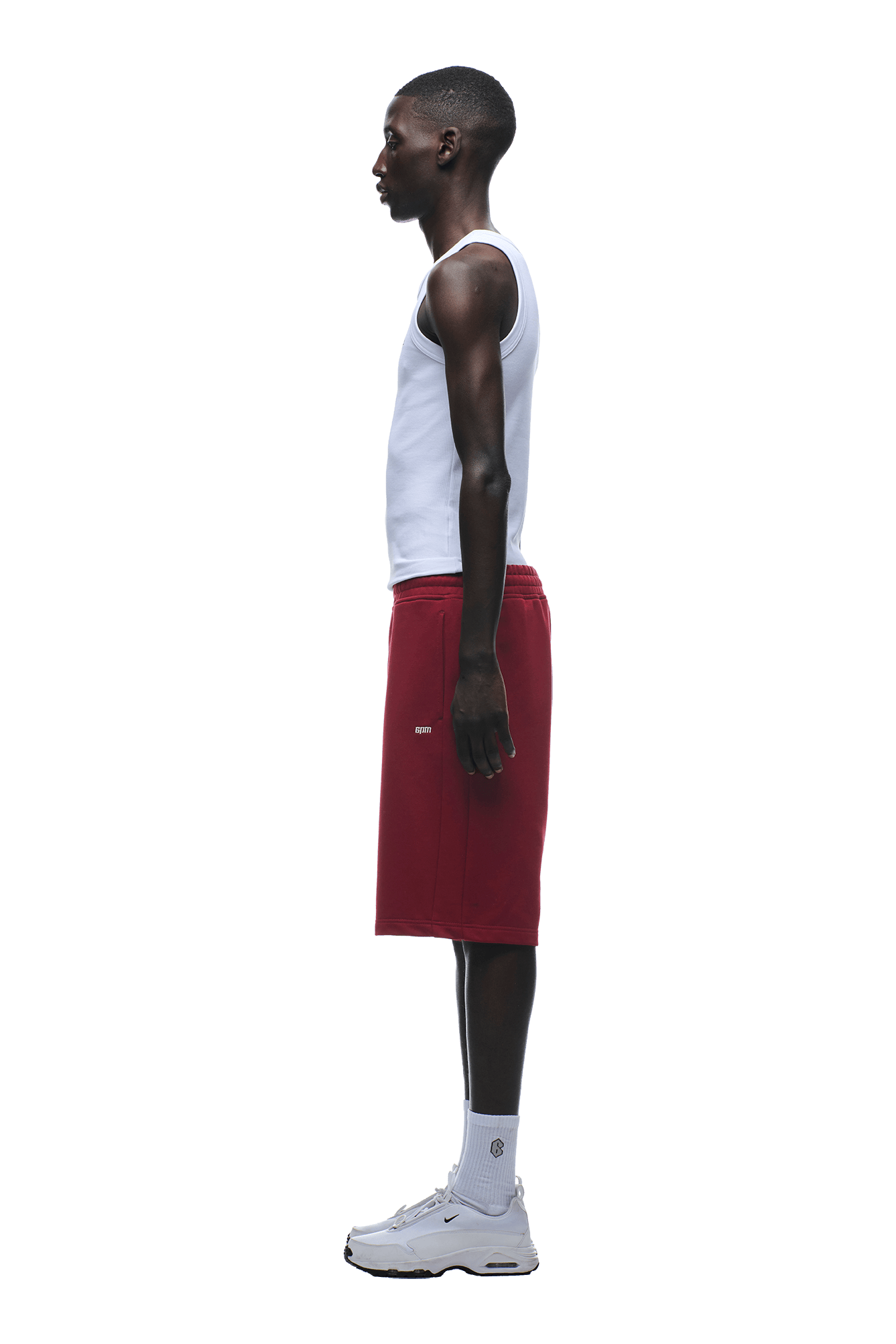 A person stands in profile facing left, wearing a white tank top, 6PM 6PM+ SHORTS WINE RED with a drawstring waist, white socks, and white sneakers against a plain background.