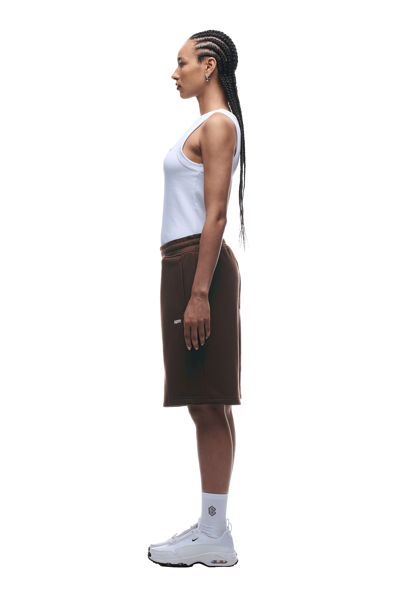 A woman in profile faces left, wearing 6PM+ SHORTS CHOCOLATE BROWN by 6PM and a white tank top. She has long braided hair, hoop earrings, white socks, and sneakers. The background is transparent.