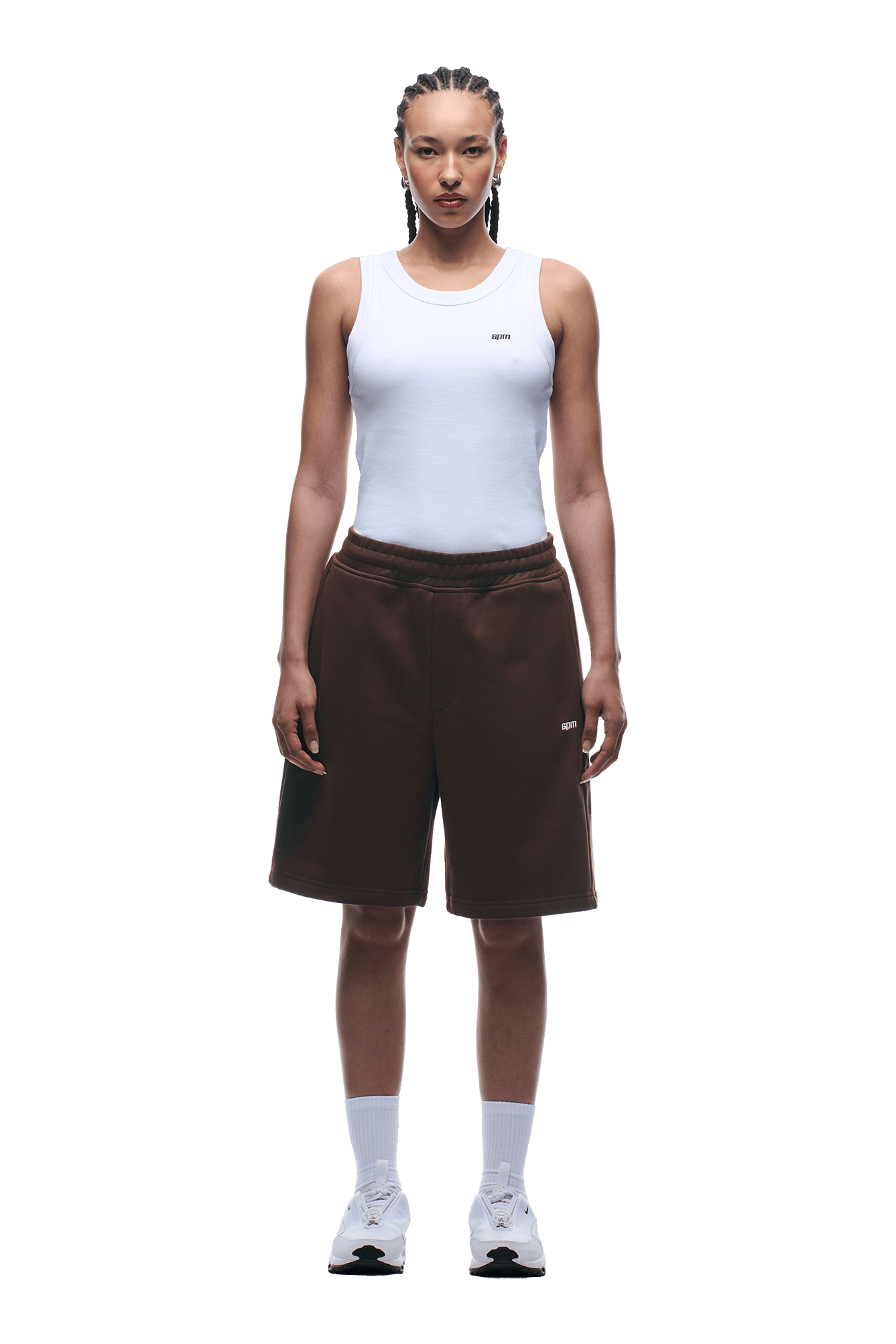 A person with braided hair wears a fitted white tank top, 6PM+ SHORTS CHOCOLATE BROWN by 6PM with a drawstring waist, white crew socks, and white athletic shoes, standing facing forward against a plain green background.