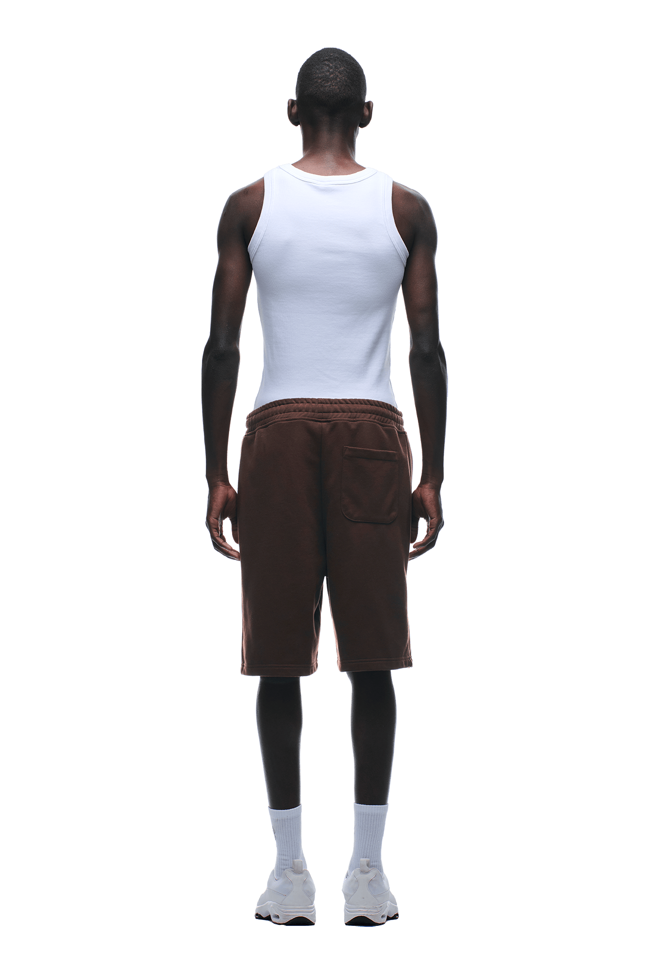A man stands facing away against a green background, wearing a white sleeveless shirt, 6PM+ SHORTS CHOCOLATE BROWN by 6PM with a drawstring waist, paired with white socks and white sneakers.