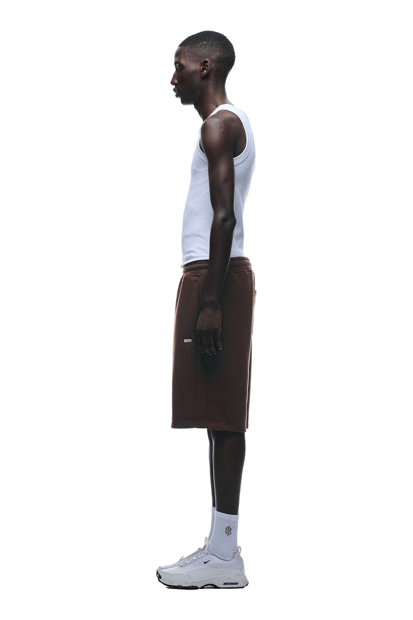 A man in profile wears the 6PM 6PM+ SHORTS CHOCOLATE BROWN, styled with a white tank top, white socks, and sneakers against a plain background.