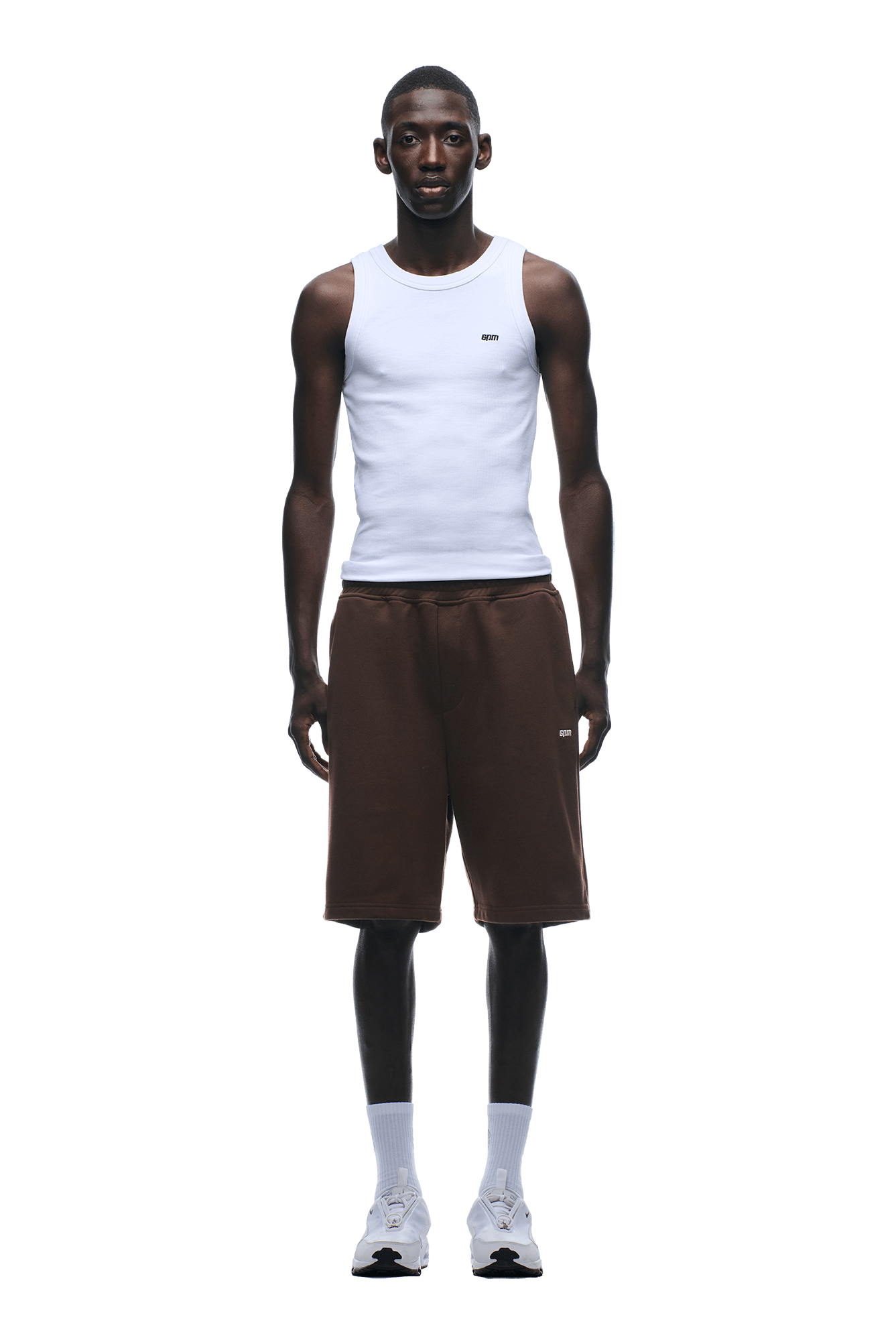 A man stands facing forward against a plain green background, wearing a white sleeveless tank top, 6PM 6PM+ SHORTS CHOCOLATE BROWN with a drawstring waist, white crew socks, and white sneakers.