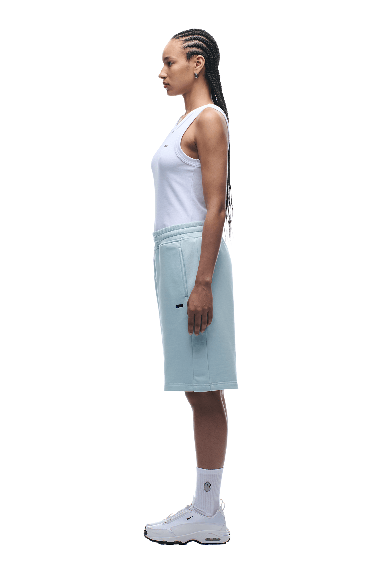 A woman with braided hair stands in profile, wearing a white tank top and 6PM+ SHORTS ICE BLUE with a drawstring waist, paired with white socks and sneakers, against a plain background.
