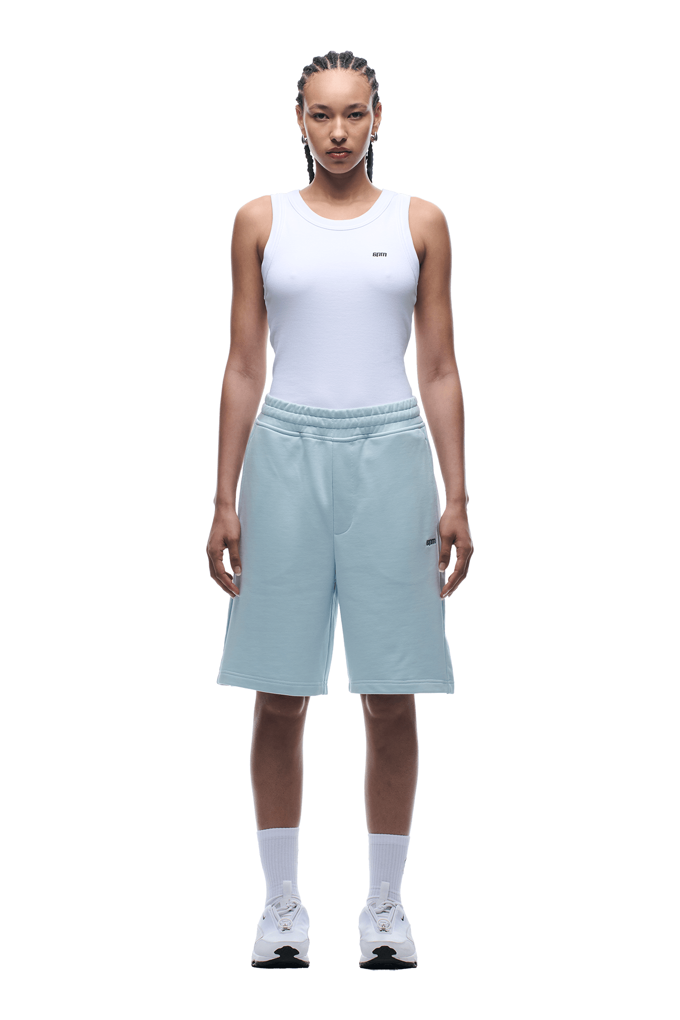 A person stands facing forward, wearing a white sleeveless top and 6PM+ SHORTS ICE BLUE with a drawstring waist, white socks, and white sneakers. Their hair is styled in cornrows against a plain green background.