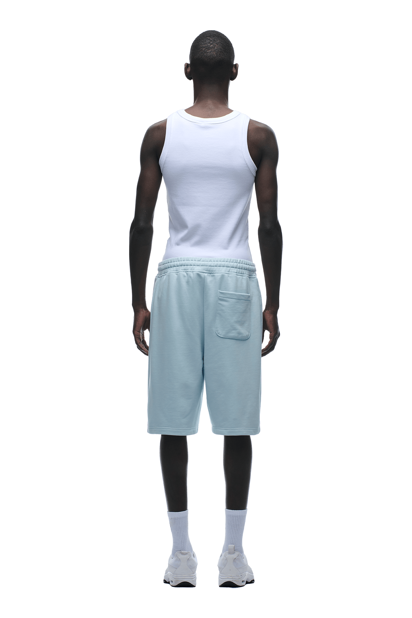 A man faces away, wearing 6PM+ SHORTS ICE BLUE—light blue, relaxed fit with a back pocket—paired with a white tank top, white crew socks, and sneakers against a plain green background.