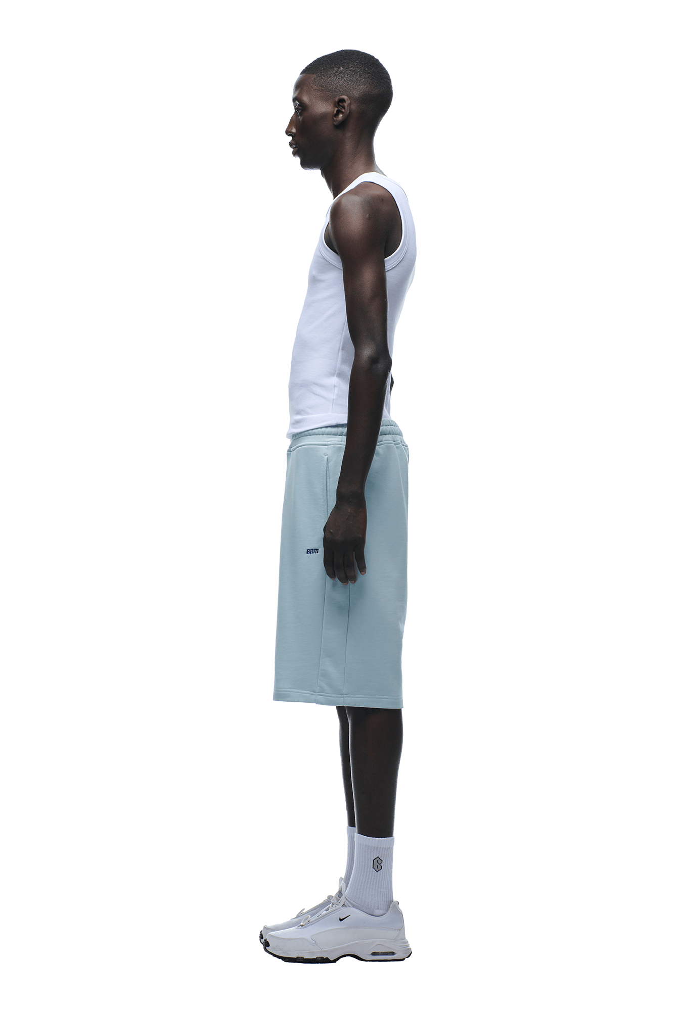 A man stands in profile wearing a relaxed fit white tank top, 6PM+ SHORTS ICE BLUE with a drawstring waist, white socks, and white sneakers against a plain background.