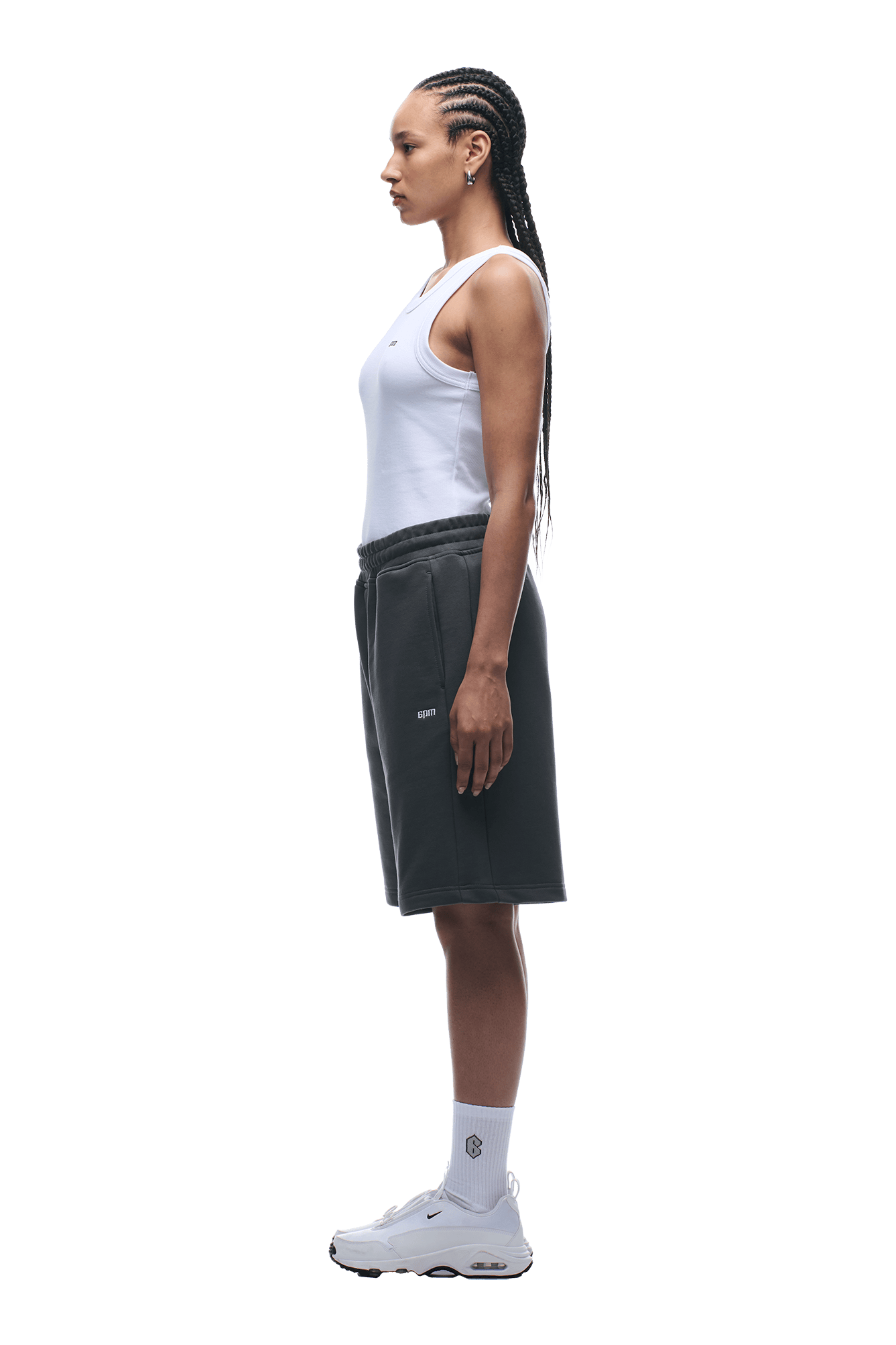 A woman with long braided hair stands in profile wearing a white tank top and 6PM+ SHORTS ANTHRACITE by 6PM, featuring a drawstring waist and relaxed knee-length fit, paired with white crew socks and sneakers on a transparent background.