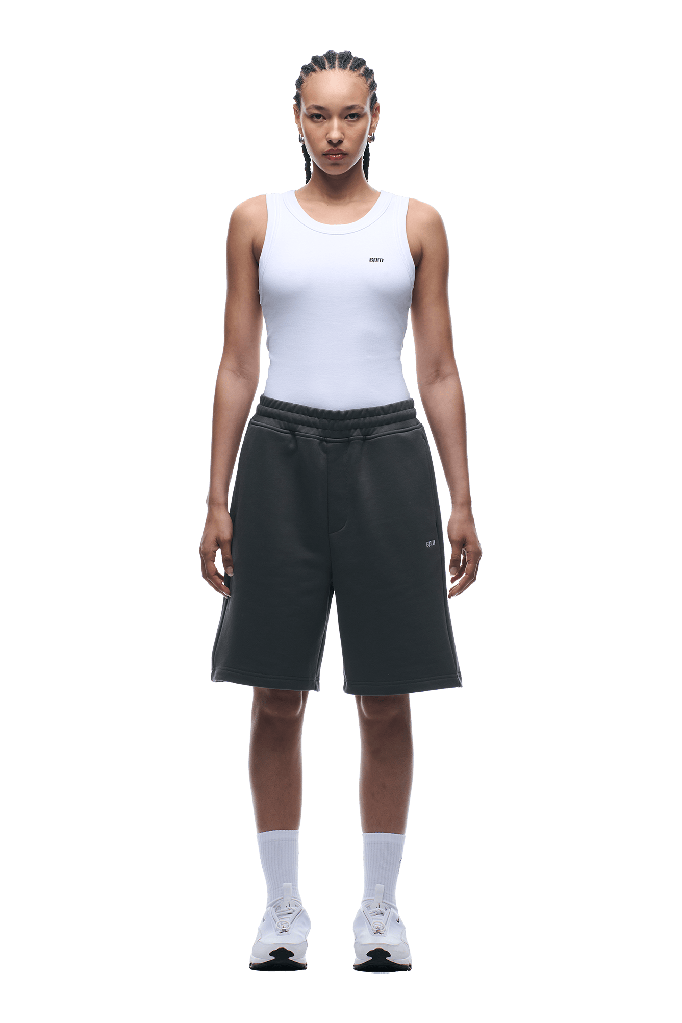 A person stands facing forward, wearing a white sleeveless top and 6PM 6PM+ SHORTS ANTHRACITE—relaxed fit, black knee-length shorts with a drawstring waist—paired with white socks and sneakers against a plain background.