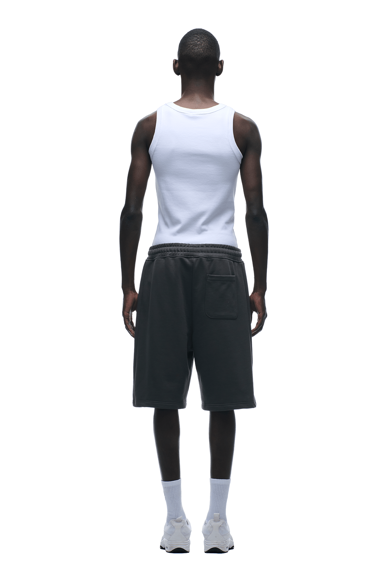 A person stands facing away, wearing a white sleeveless tank top and 6PM+ SHORTS ANTHRACITE by 6PM—dark, knee-length shorts with a drawstring waist—paired with white socks and sneakers against a plain background.