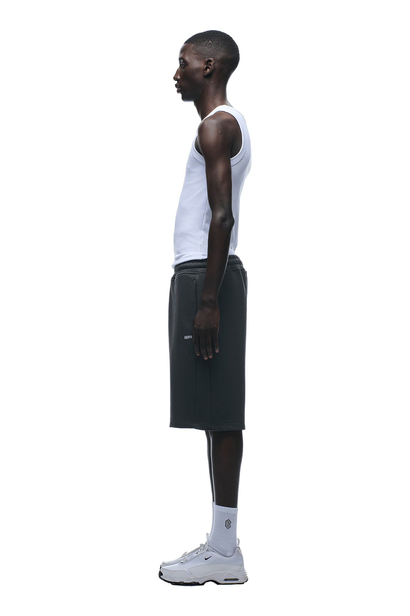 A man stands in profile, facing left, wearing a white tank top and 6PM+ SHORTS ANTHRACITE by 6PM—dark, relaxed-fit knee-length shorts—paired with white socks and sneakers, set against a plain background.