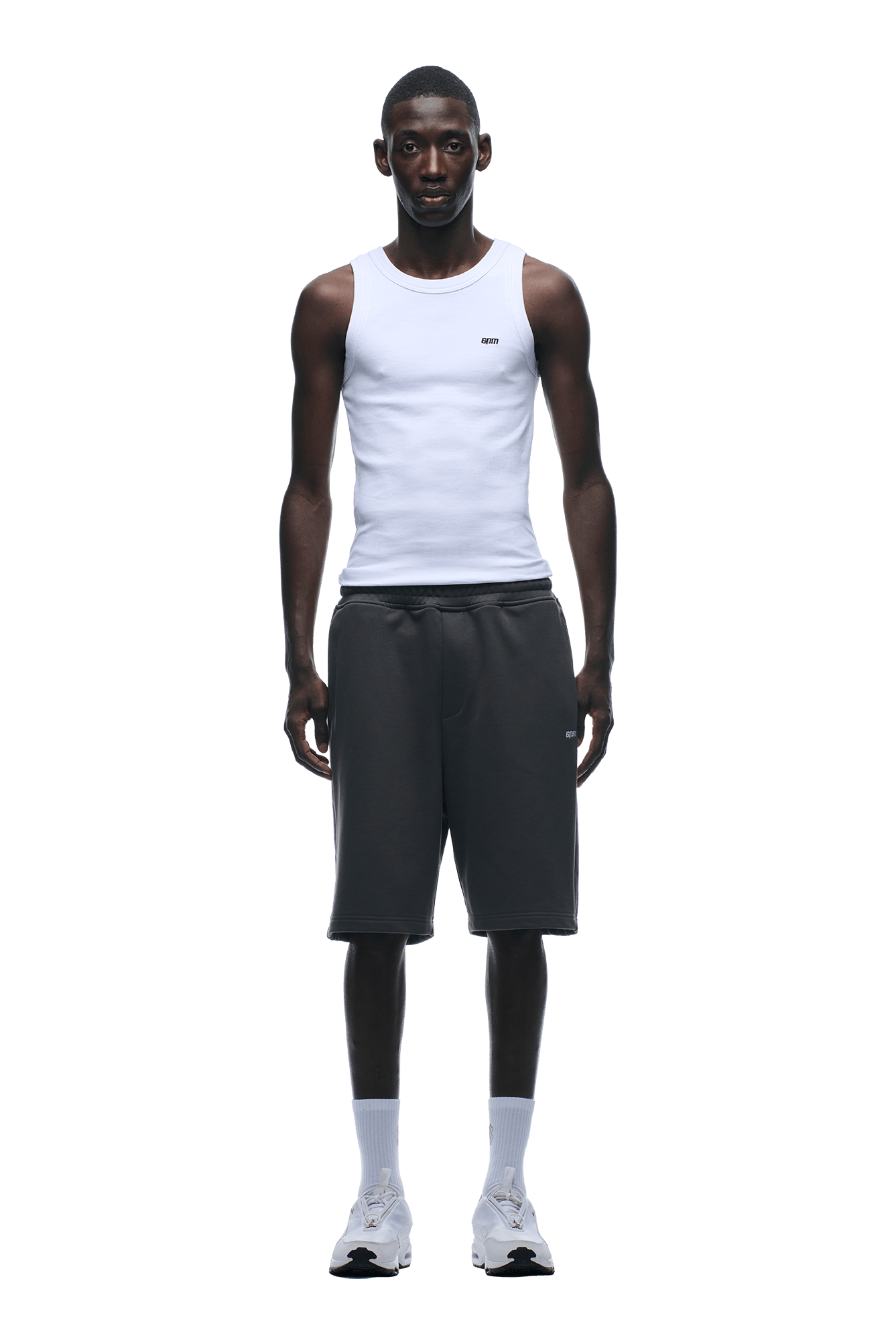 A man faces forward, wearing a white sleeveless tank top and 6PM+ SHORTS ANTHRACITE by 6PM—dark knee-length shorts with a relaxed fit—paired with white crew socks and athletic shoes, set against a plain background.