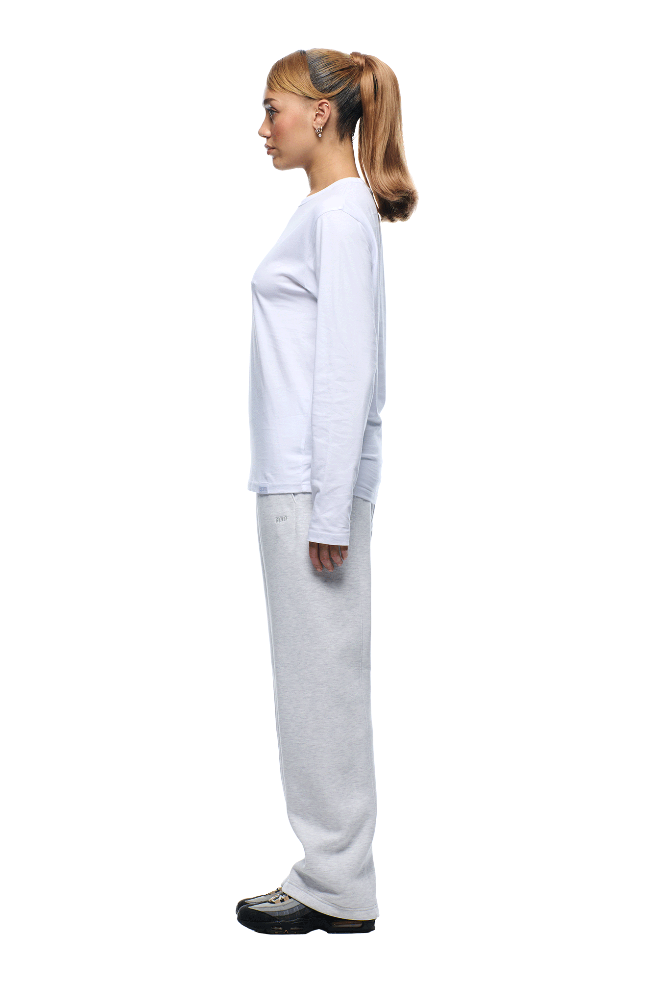 A woman with light brown hair in a high ponytail stands in profile, wearing the 6PM+ SLIM FIT LONGSLEEVE WHITE by 6PM, paired with light gray sweatpants and black sneakers against a minimalist white background.