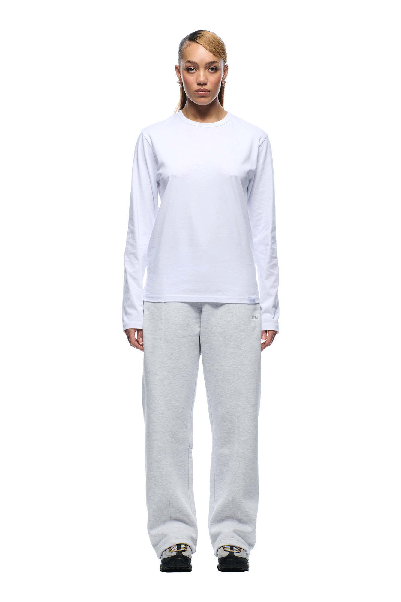 A person stands facing forward, wearing the 6PM+ SLIM FIT LONGSLEEVE WHITE by 6PM and light gray sweatpants with black sneakers. The minimalist style is accentuated by the plain white background.