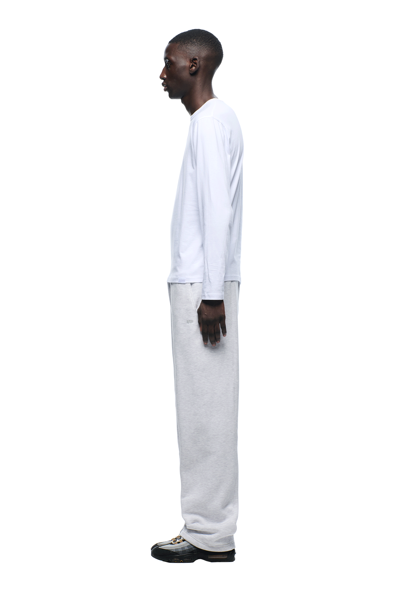 A man stands in profile against a white background, wearing the 6PM 6PM+ SLIM FIT LONGSLEEVE WHITE shirt, light gray sweatpants, and black sneakers with white soles.