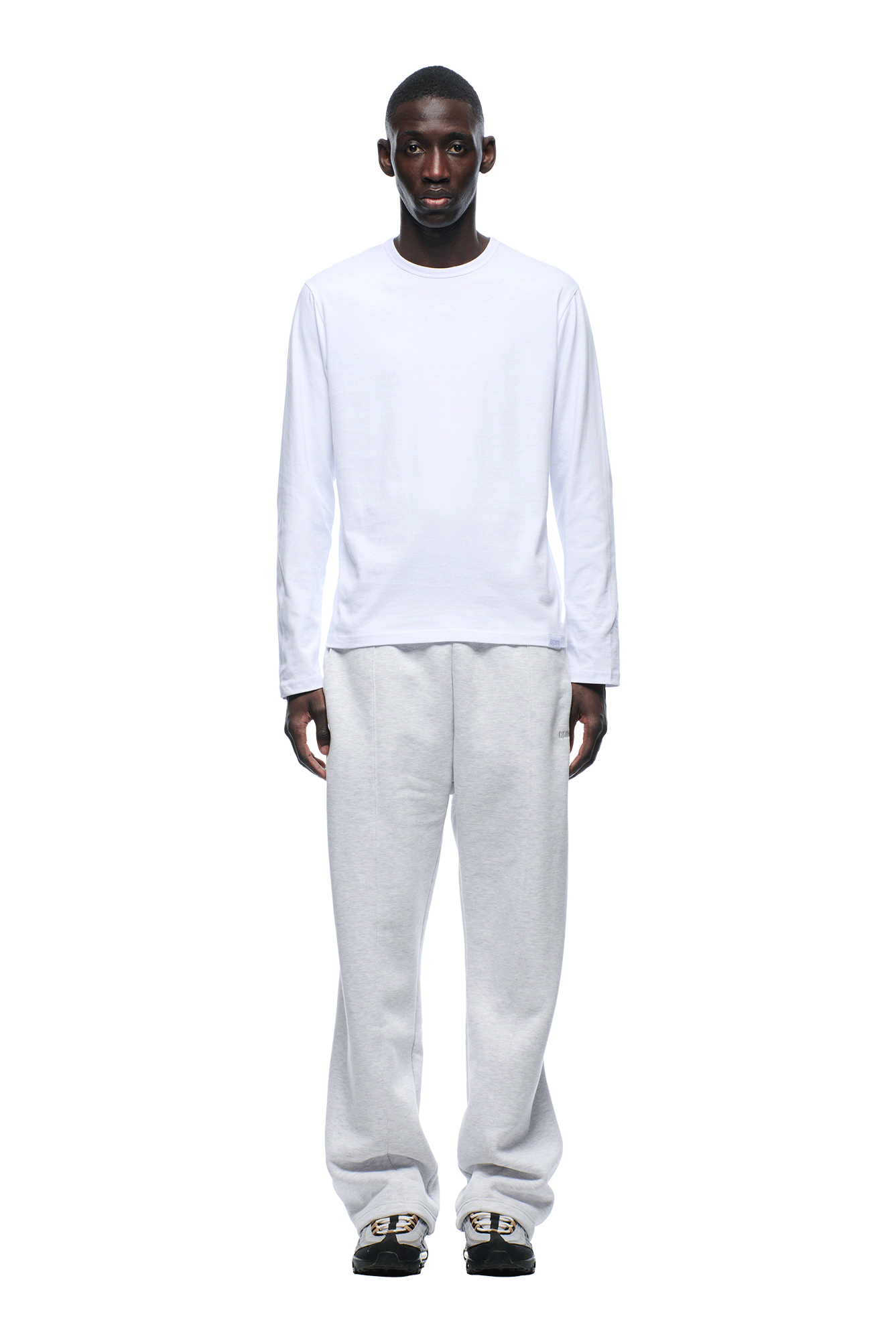 A man faces forward against a white background, wearing the 6PM+ SLIM FIT LONGSLEEVE WHITE by 6PM, light gray sweatpants, and black slide sandals with gold chain detailing.