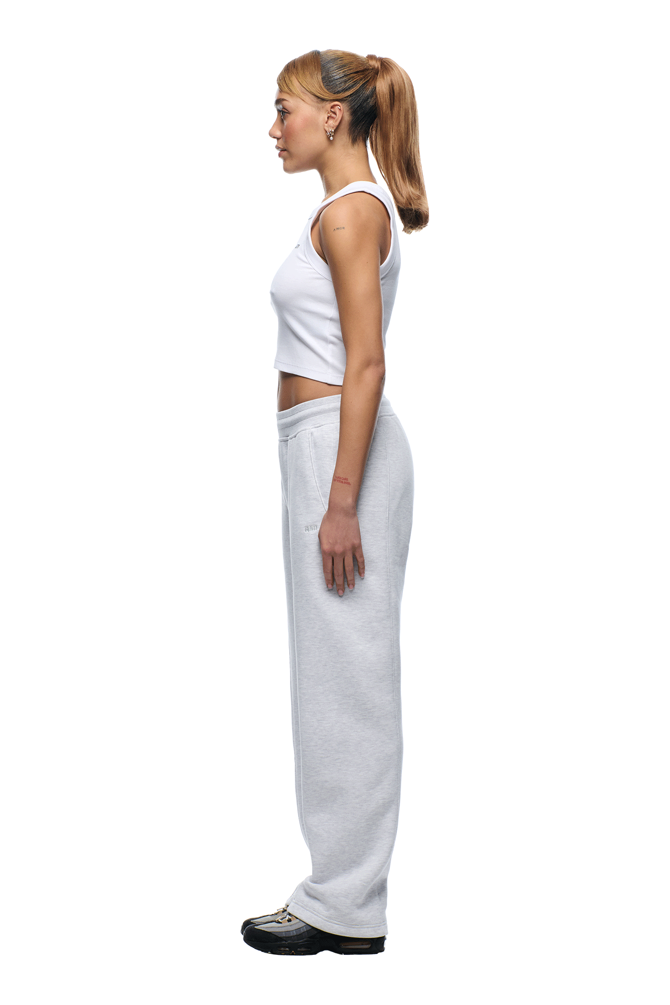 A young woman with a ponytail stands in profile against a white background, wearing a white sleeveless crop top, black sneakers, and 6PM+ SLIM FIT JOGGER LIGHT GREY sweatpants by 6PM.