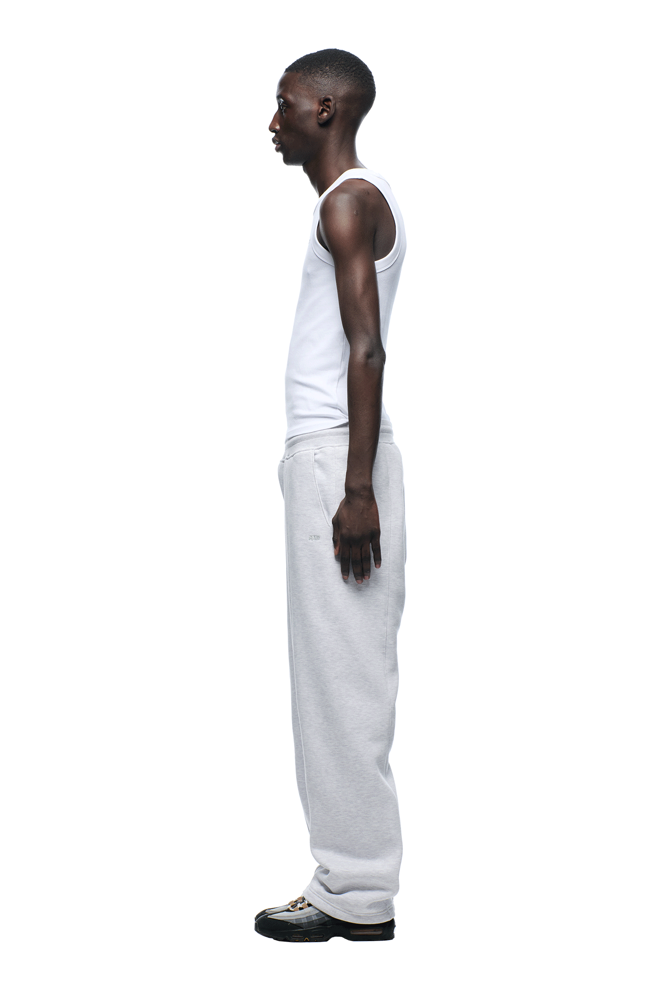 A man stands in profile against a white background, wearing a heavyweight white tank top, 6PM+ Slim Fit Jogger Light Grey sweatpants by 6PM, and black sneakers. His arms rest by his sides as he looks straight ahead.