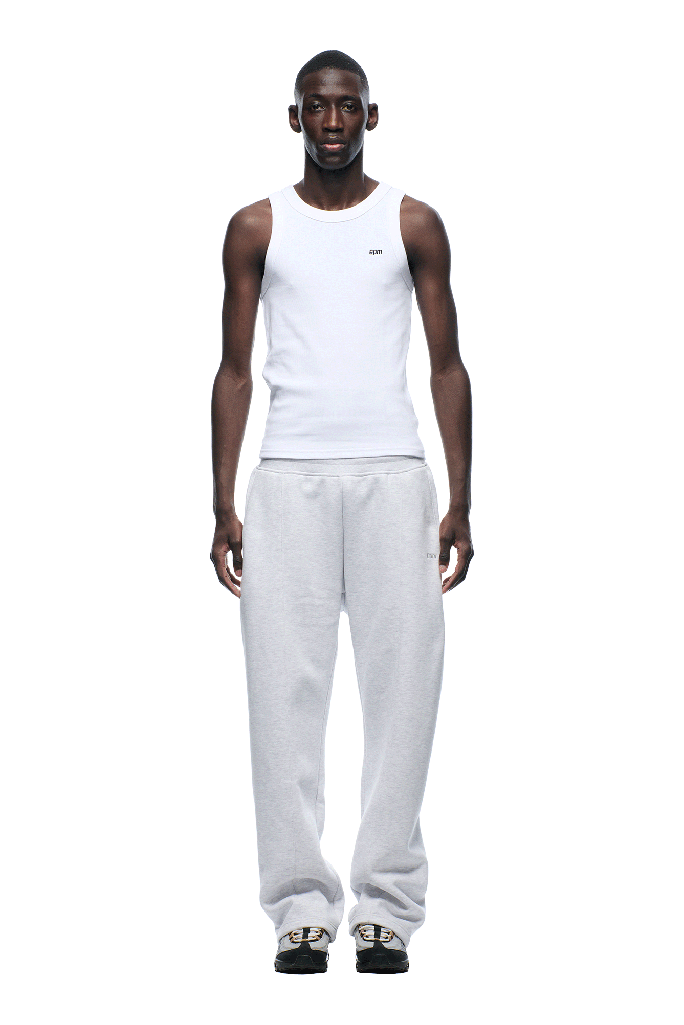 A man faces forward against a plain white background, wearing a white sleeveless tank top and 6PM 6PM+ SLIM FIT JOGGER LIGHT GREY sweatpants, paired with black slip-on shoes featuring gold details.