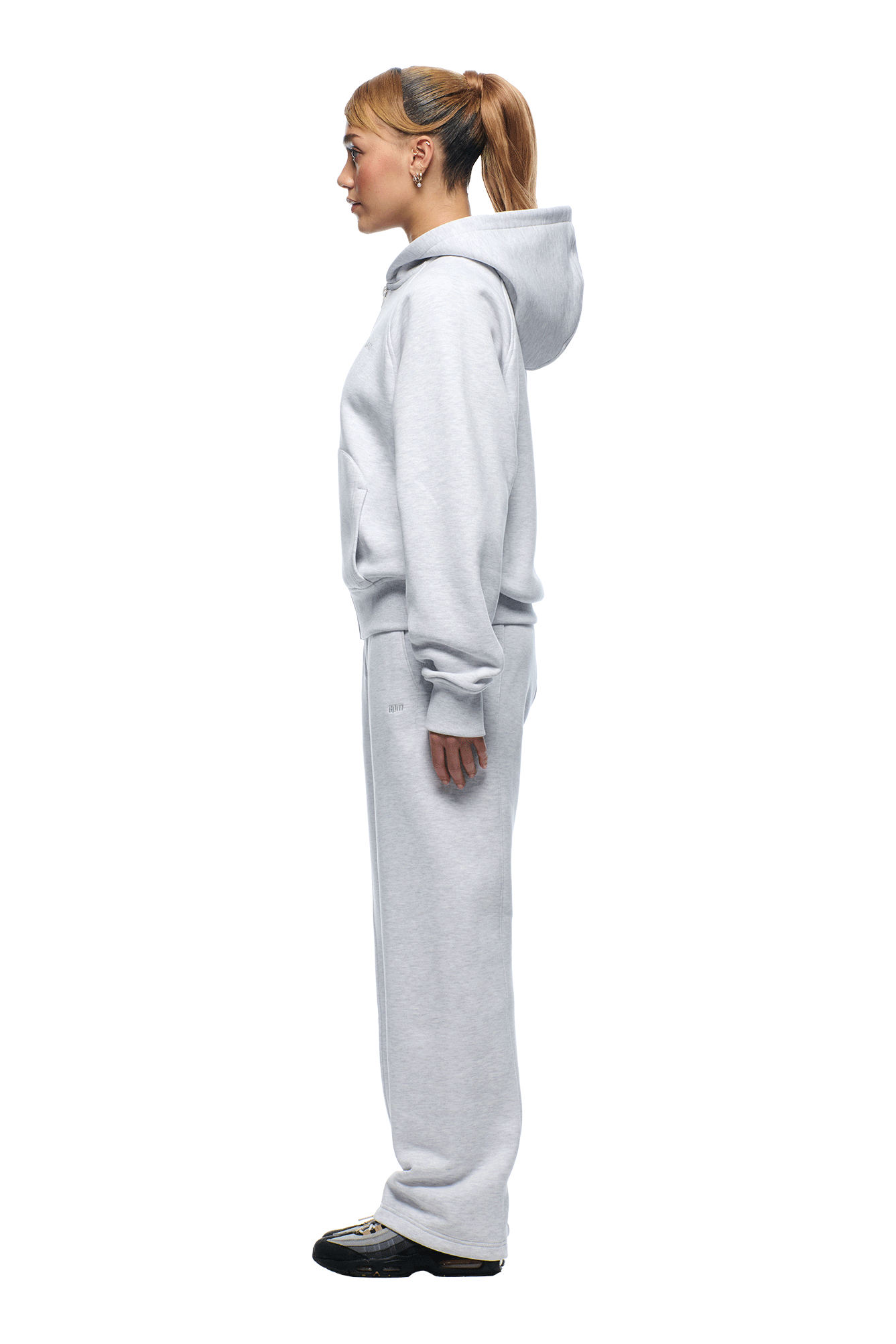 A woman in profile faces left, wearing the 6PM 6PM+ SLIM ZIP HOODIE LIGHT GREY and matching sweatpants with black sneakers. She has a ponytail and stands against a plain white background, highlighting the clean finish.