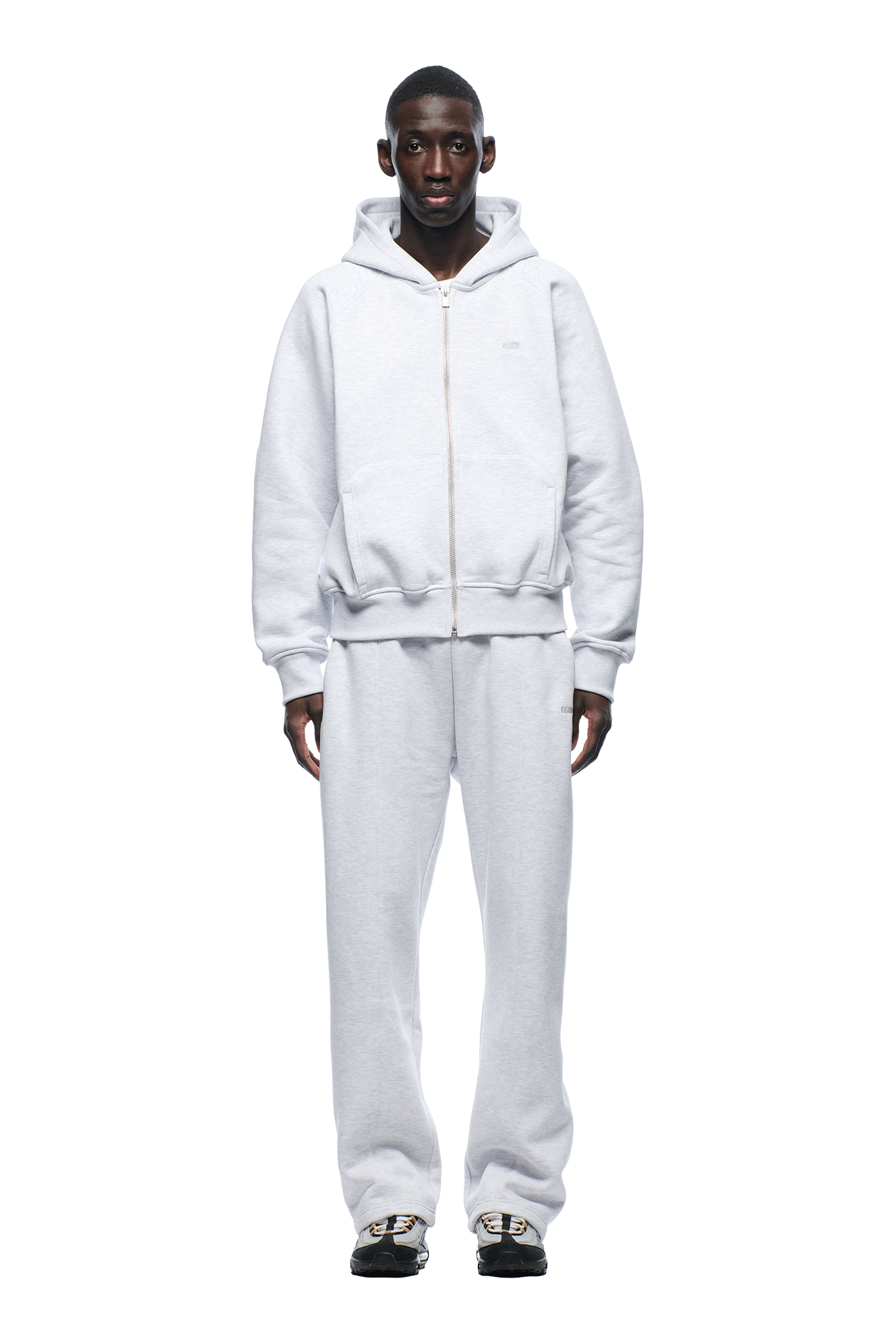 A person stands facing forward, wearing the 6PM+ SLIM ZIP HOODIE LIGHT GREY by 6PM with matching sweatpants and black and white sneakers, giving a clean, structured look against a plain white background.