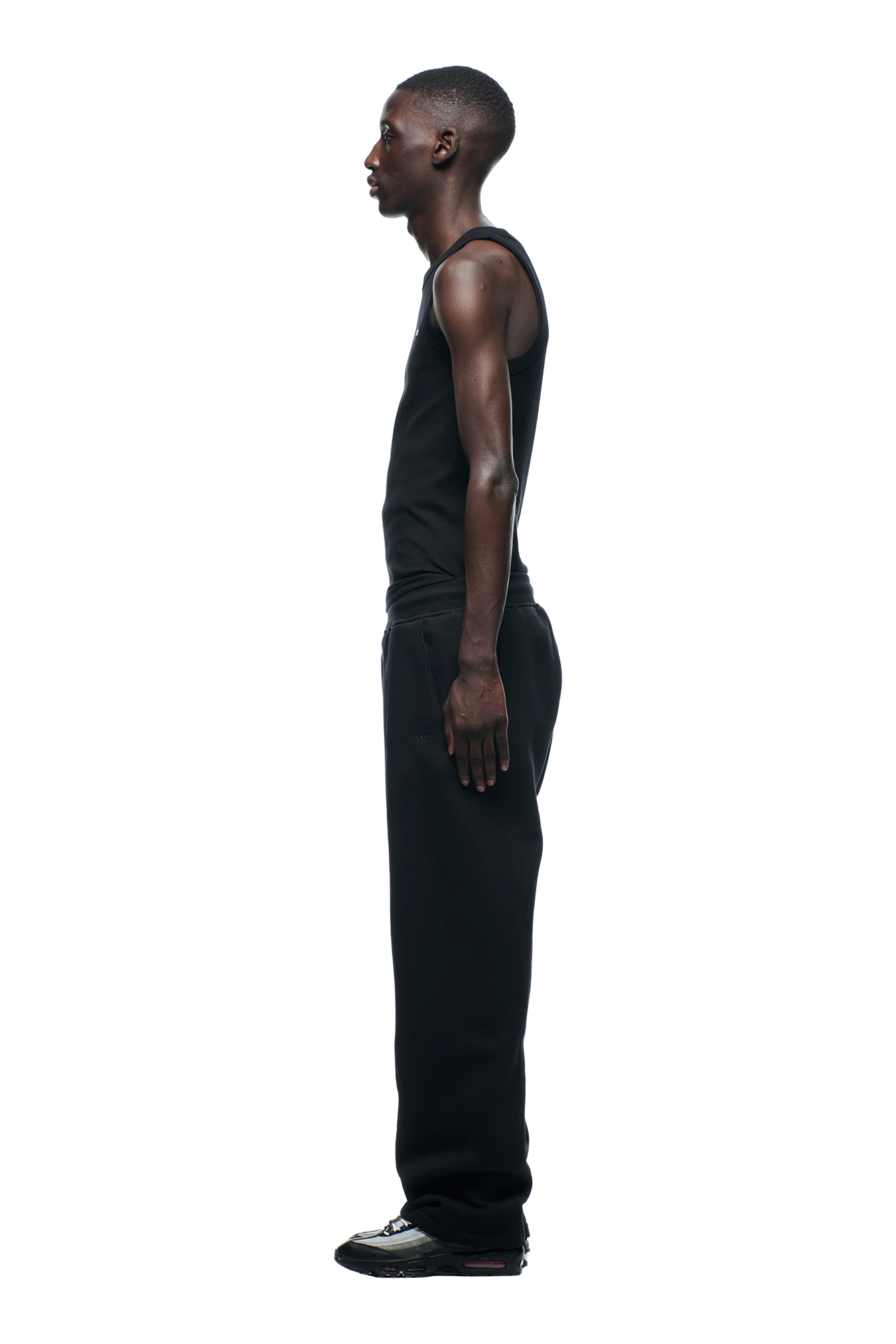 A man in profile, facing left, wears a black tank top and 6PM+ Slim Fit Jogger Faded Black pants by 6PM with black sneakers, set against a white background.