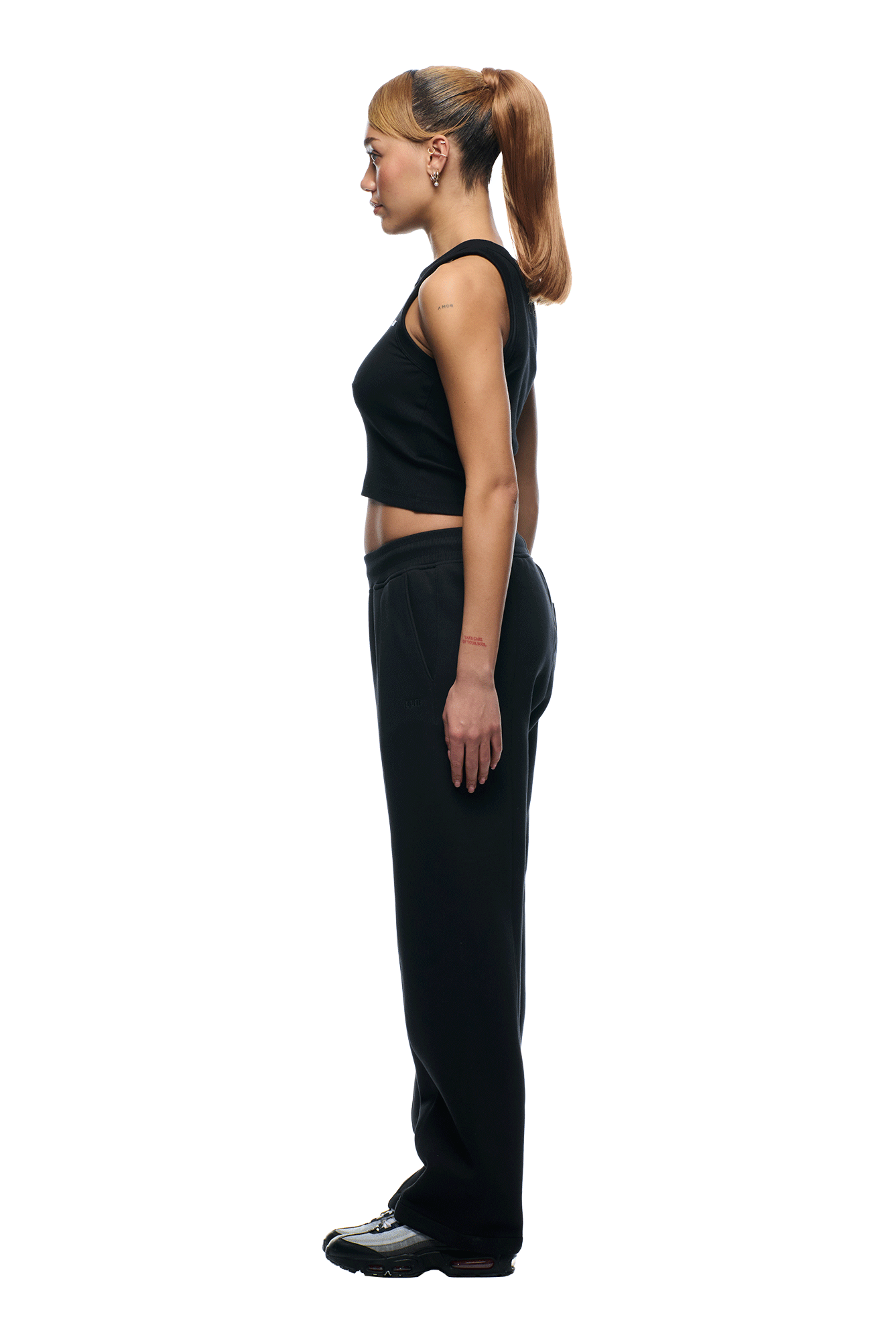 A woman with light brown hair in a ponytail wears a black sleeveless top and 6PM+ SLIM FIT JOGGER FADED BLACK by 6PM, paired with black sneakers, standing in profile against a white background.