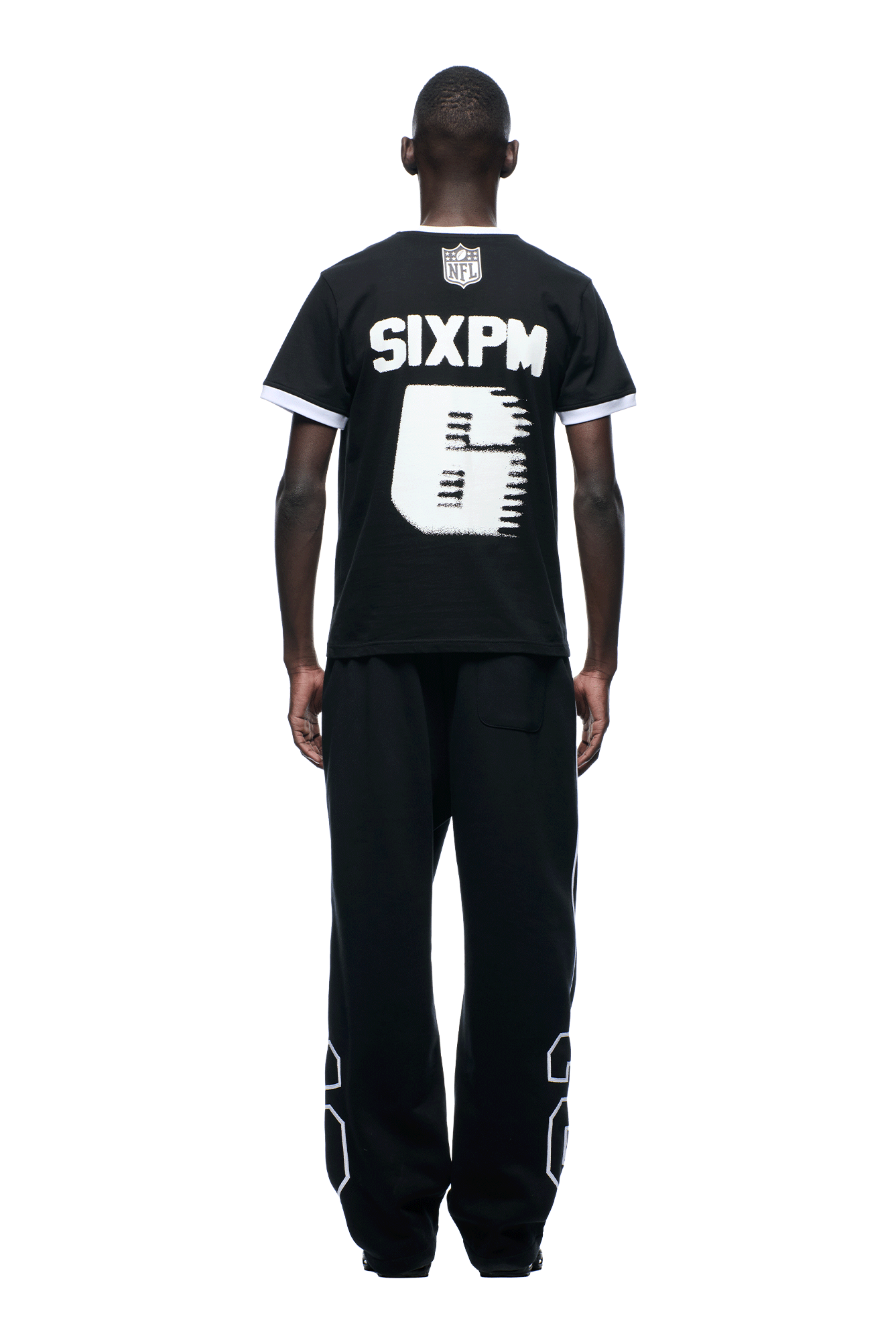 A person faces away, wearing the 6PM NFL BLURRED T-SHIRT: a black tee with bold white SIXPM text and a large, partially blurred number 5, paired with black pants featuring big white numbers. The background is plain white.