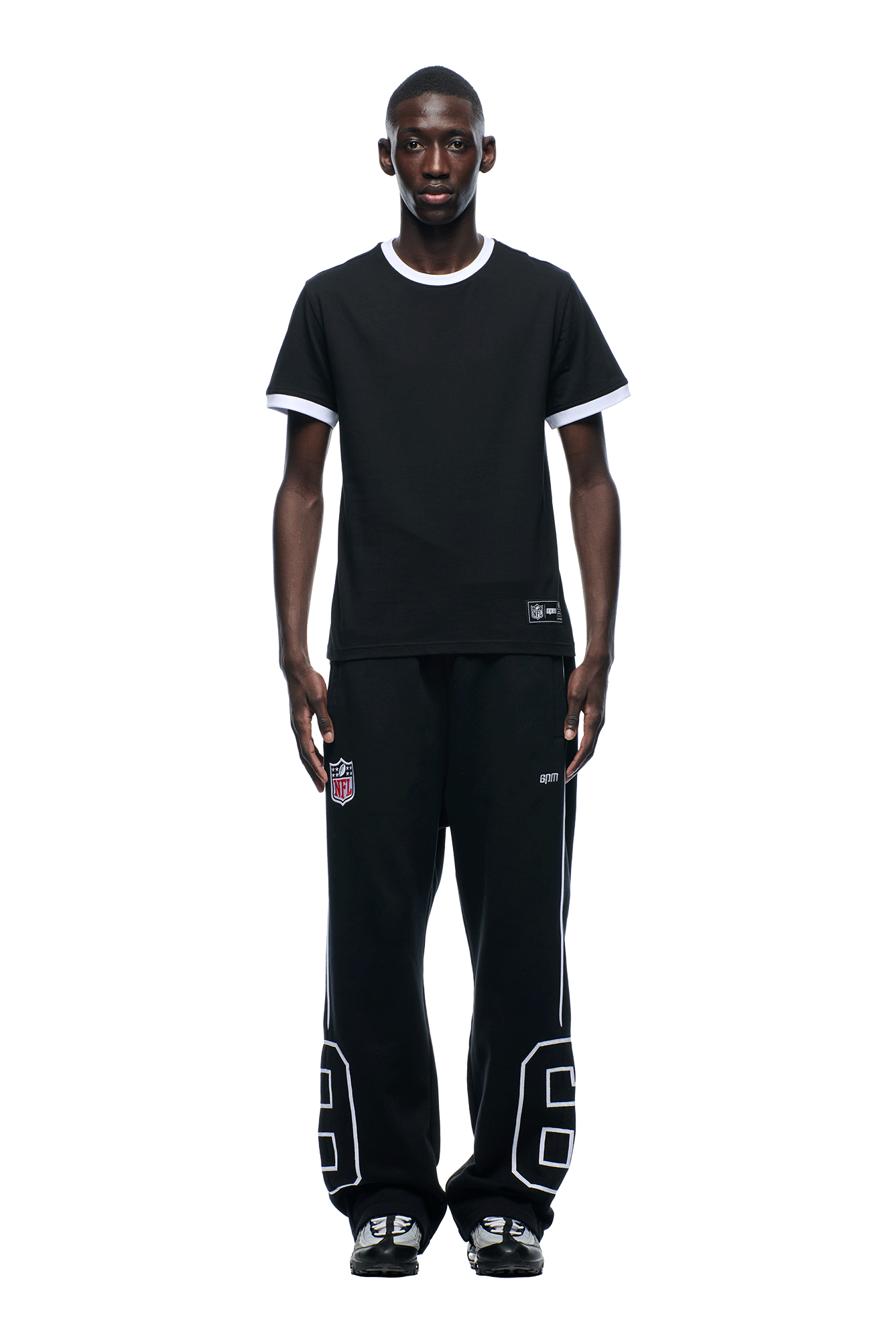 A man stands facing forward against a white background, wearing the 6PM NFL BLURRED T-SHIRT with white trim, black pants featuring large white "92" numbers, and black sneakers.