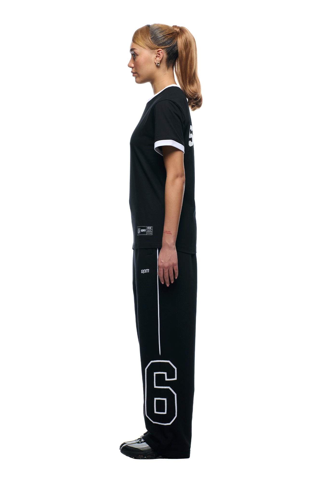 A woman with light brown hair in a ponytail stands in profile, wearing the 6PM NFL BLURRED T-SHIRT and black sweatpants with a large white number 6 on the left leg, set against a plain white background.