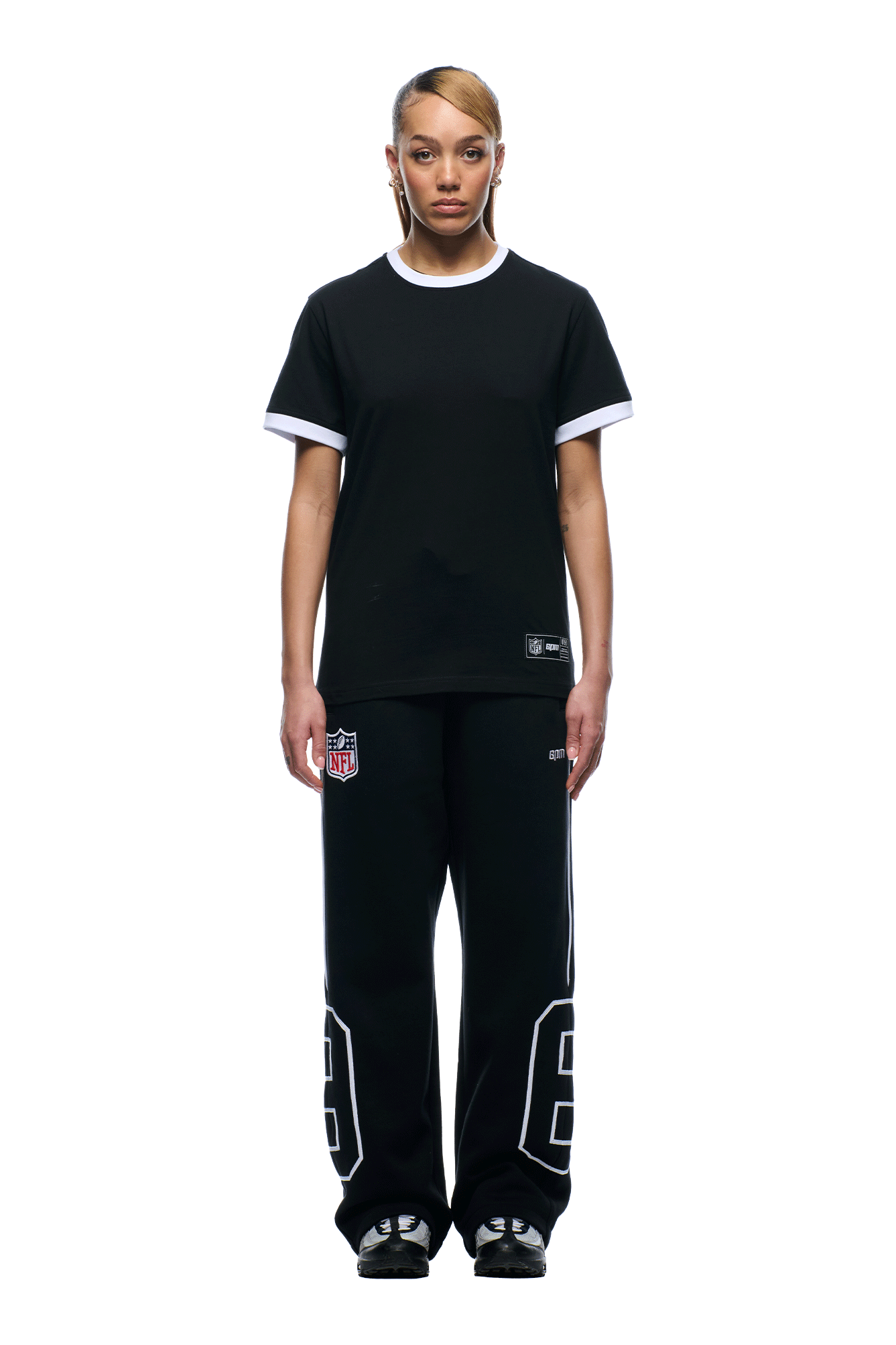 A person with short, side-parted hair and neutral expression stands facing forward against a white background, wearing 6PM's NFL BLURRED T-SHIRT and black sweatpants with white numbers and an NFL logo.