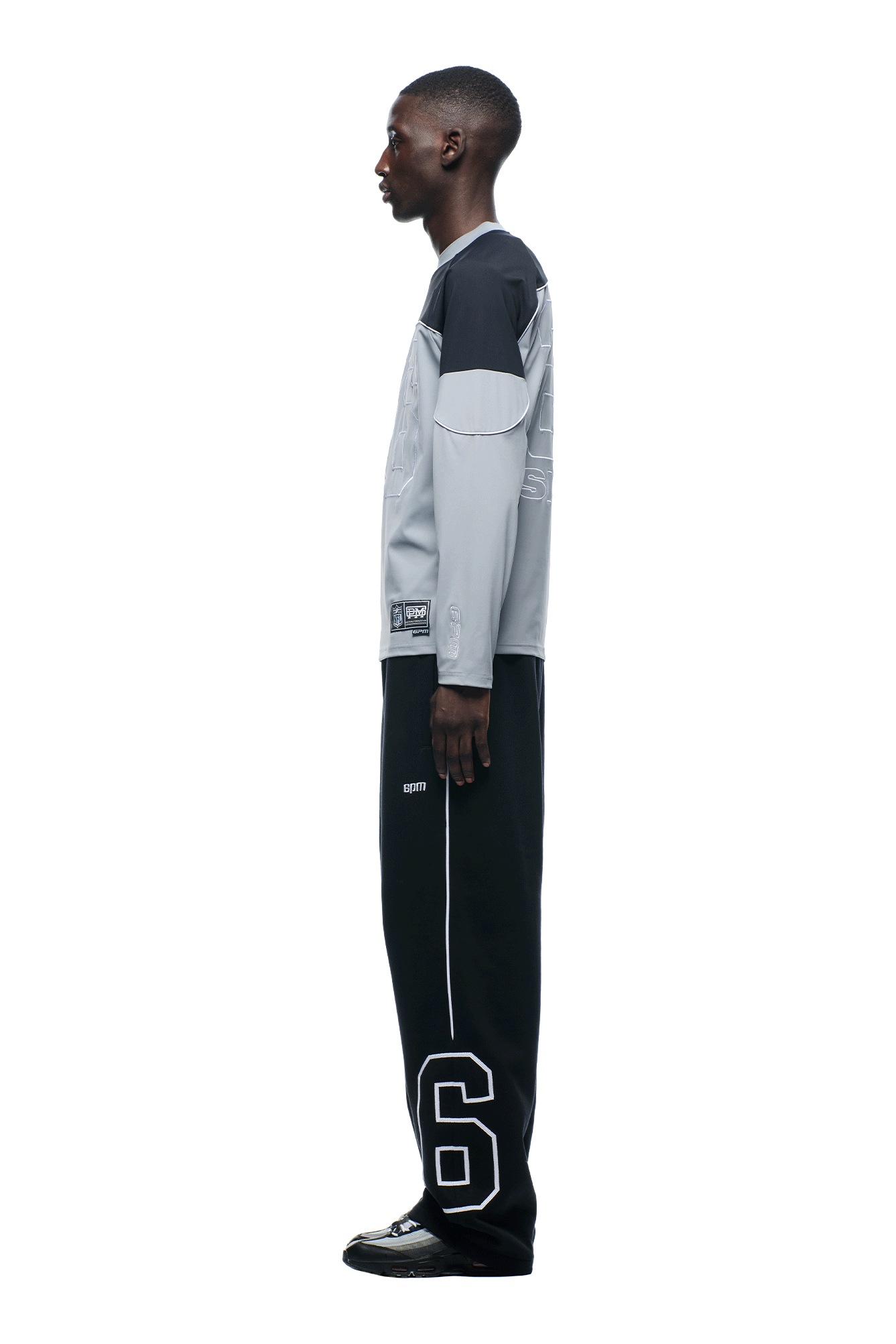 A person stands in profile against a plain background, wearing the 6PM NFL SCUBA LONGSLEEVE GREY shirt and black pants with a large white number 6 on the lower leg, paired with black shoes.