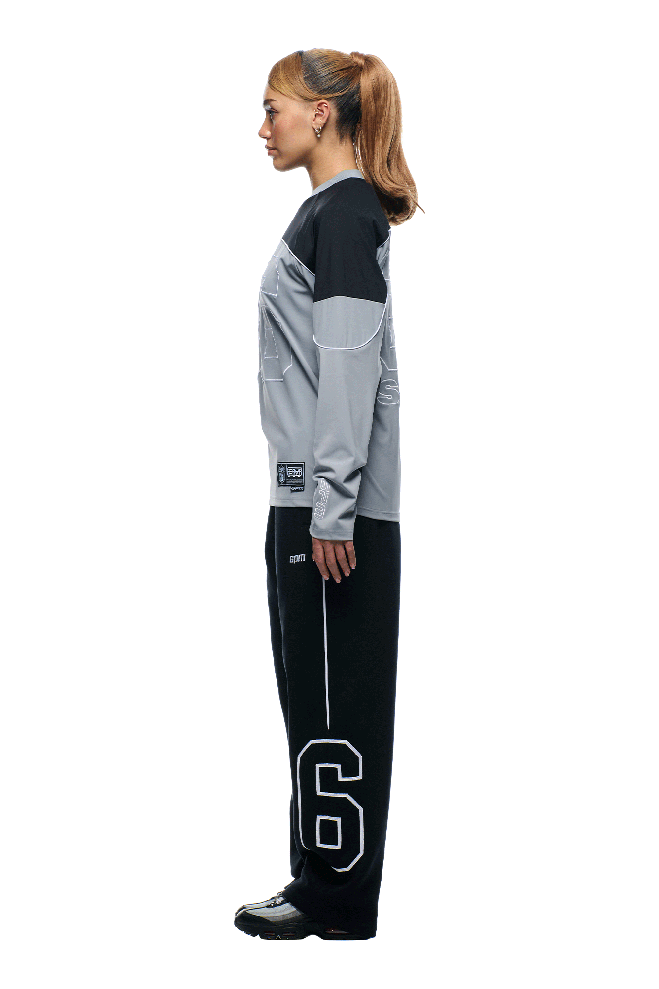 A woman with a ponytail stands in profile against a white background, wearing the 6PM NFL SCUBA LONGSLEEVE GREY shirt and black pants with a large white number 6 on the left leg.