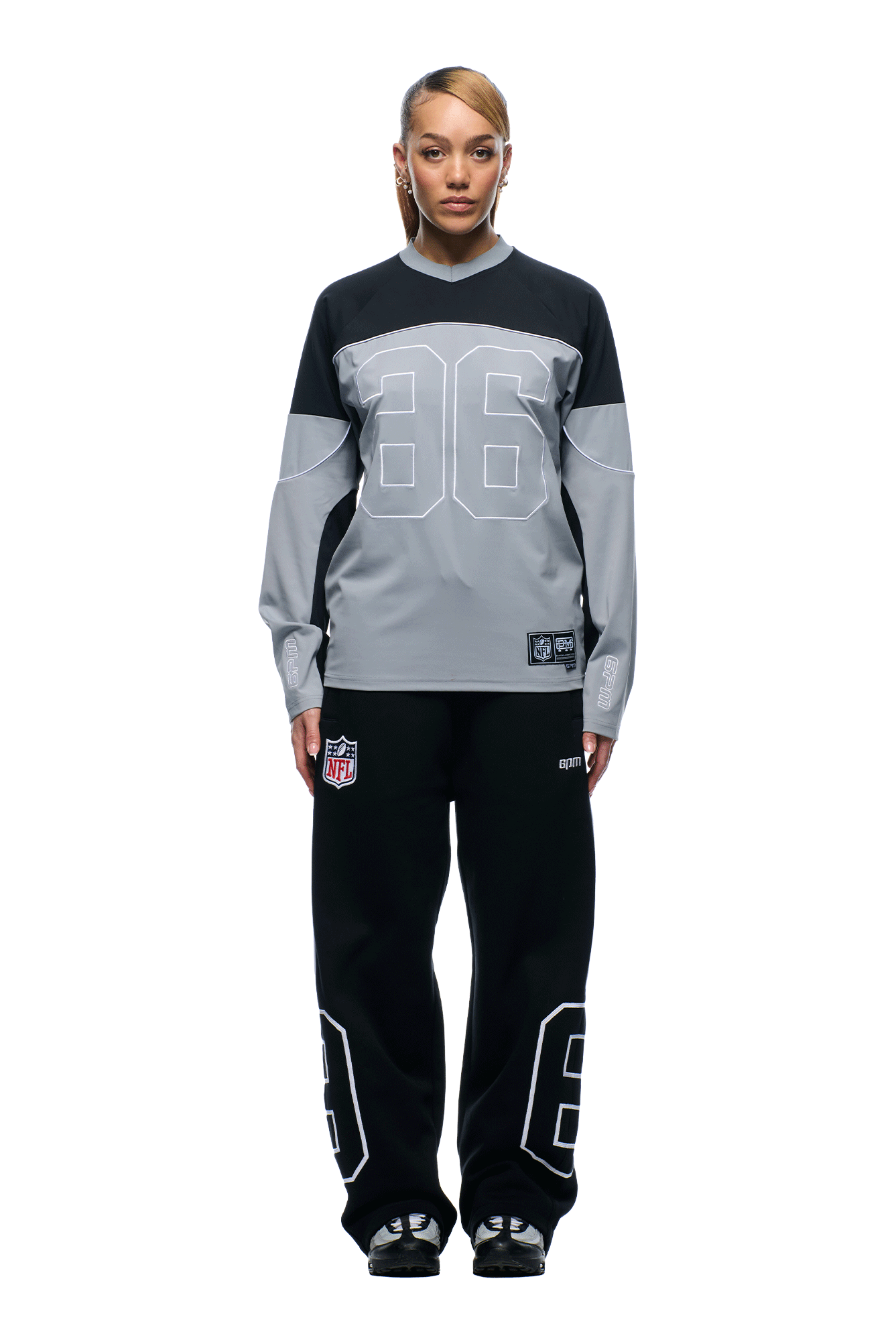 A woman with side-swept hair wears the 6PM NFL SCUBA LONGSLEEVE GREY top with "86" on the front, plus black pants with "86" and an NFL logo patch, and black shoes. She stands facing forward against a white background.