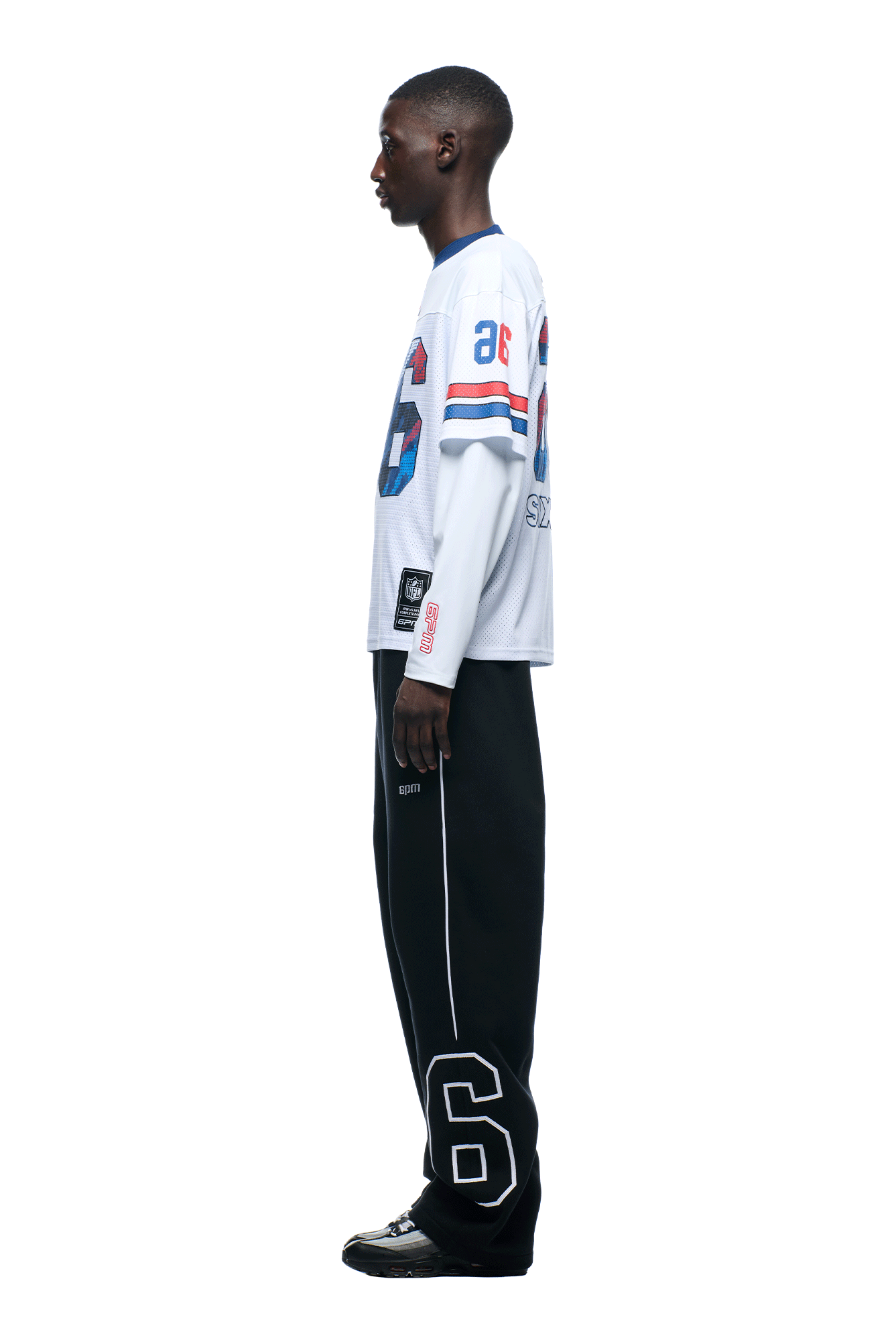 A person in profile stands against a plain background, wearing the 6PM NFL Layered Mesh Longsleeve White jersey with blue and red accents, black pants with a large white number 6 on the lower leg, and black sneakers.