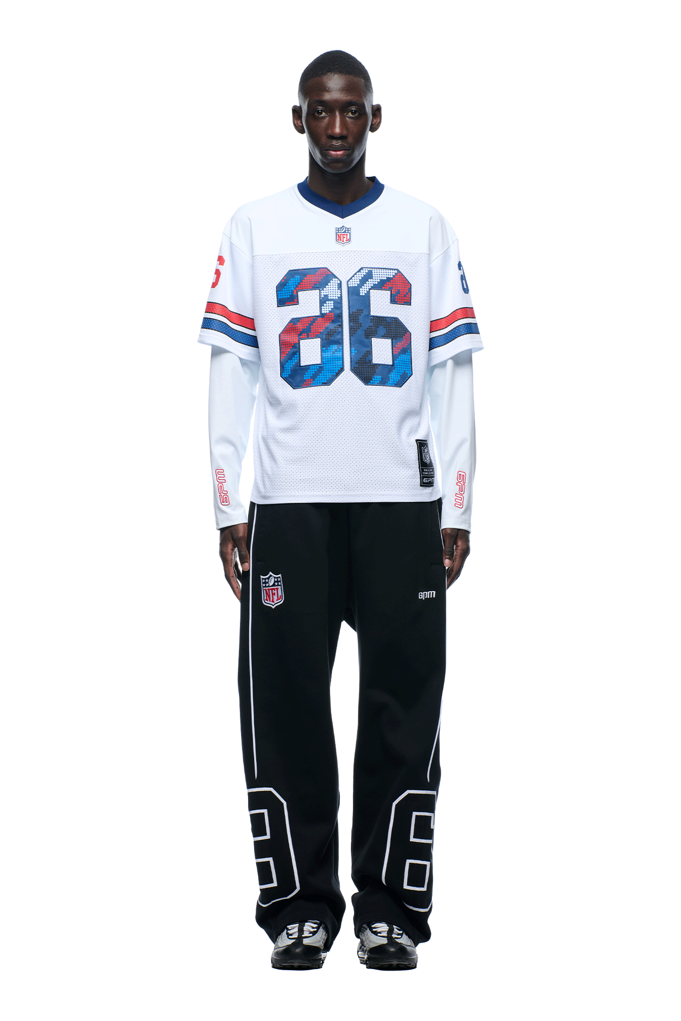 A man with short hair is wearing the 6PM NFL LAYERED MESH LONGSLEEVE WHITE jersey with blue and red stripes and the number 26, paired with black pants featuring white NFL logos and the number 26, and black sneakers.