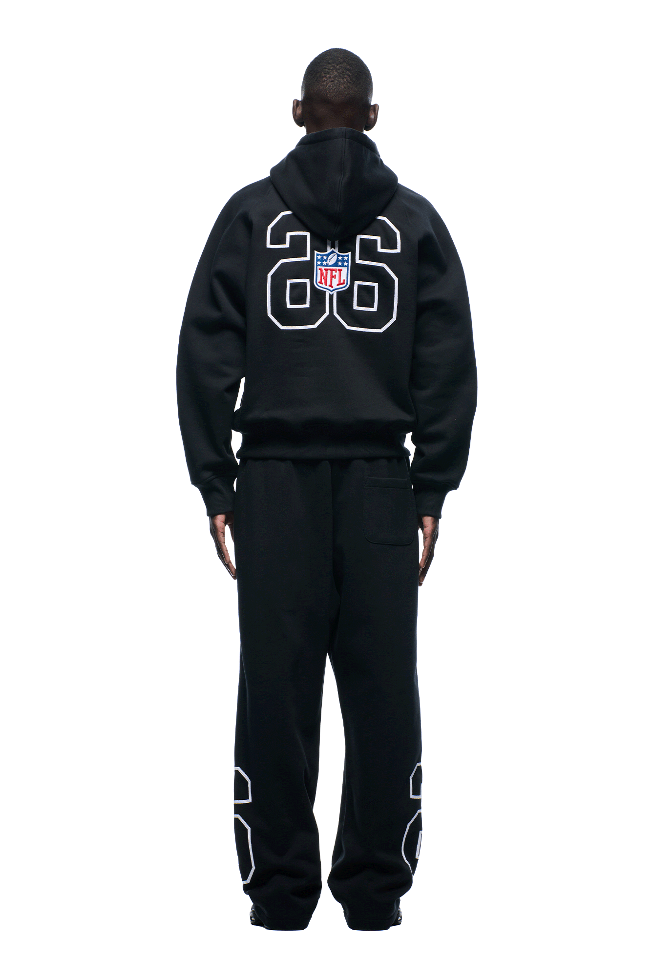 A person wearing a black 6PM NFL PIPING HOODIE with a large white 26 and NFL logo on the back, plus matching sweatpants with the number 26 on the lower legs, stands against a white background.