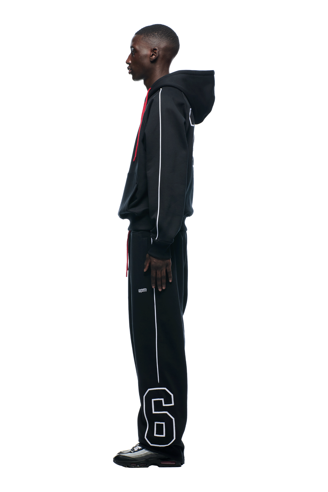 A man stands in profile against a white background wearing the 6PM NFL PIPING HOODIE and matching track pants with white piping and a large number 6 on the left leg, paired with black shoes.