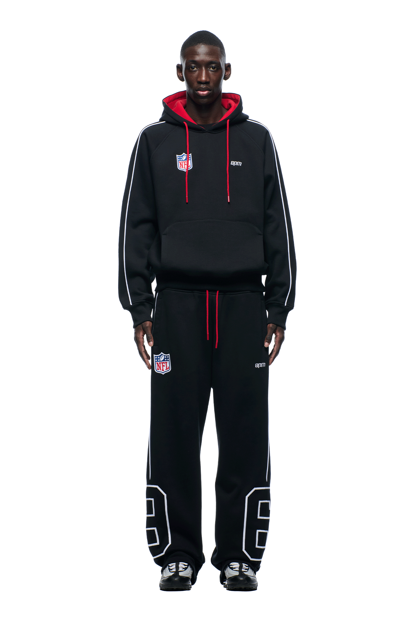 A person faces forward wearing the 6PM NFL PIPING HOODIE with red lining, matching pants, and black sneakers. Both hoodie and pants feature NFL patches and white accents, set against a plain white background.