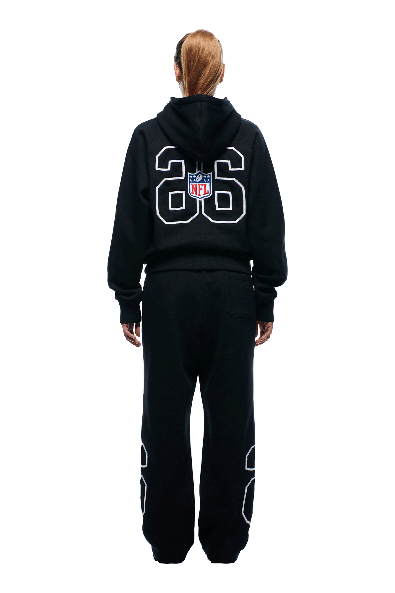 A person with long hair in a ponytail wears a black 6PM NFL PIPING HOODIE and matching sweatpants with the number 26 and NFL logo on the back, standing against a plain white background, facing away from the camera.