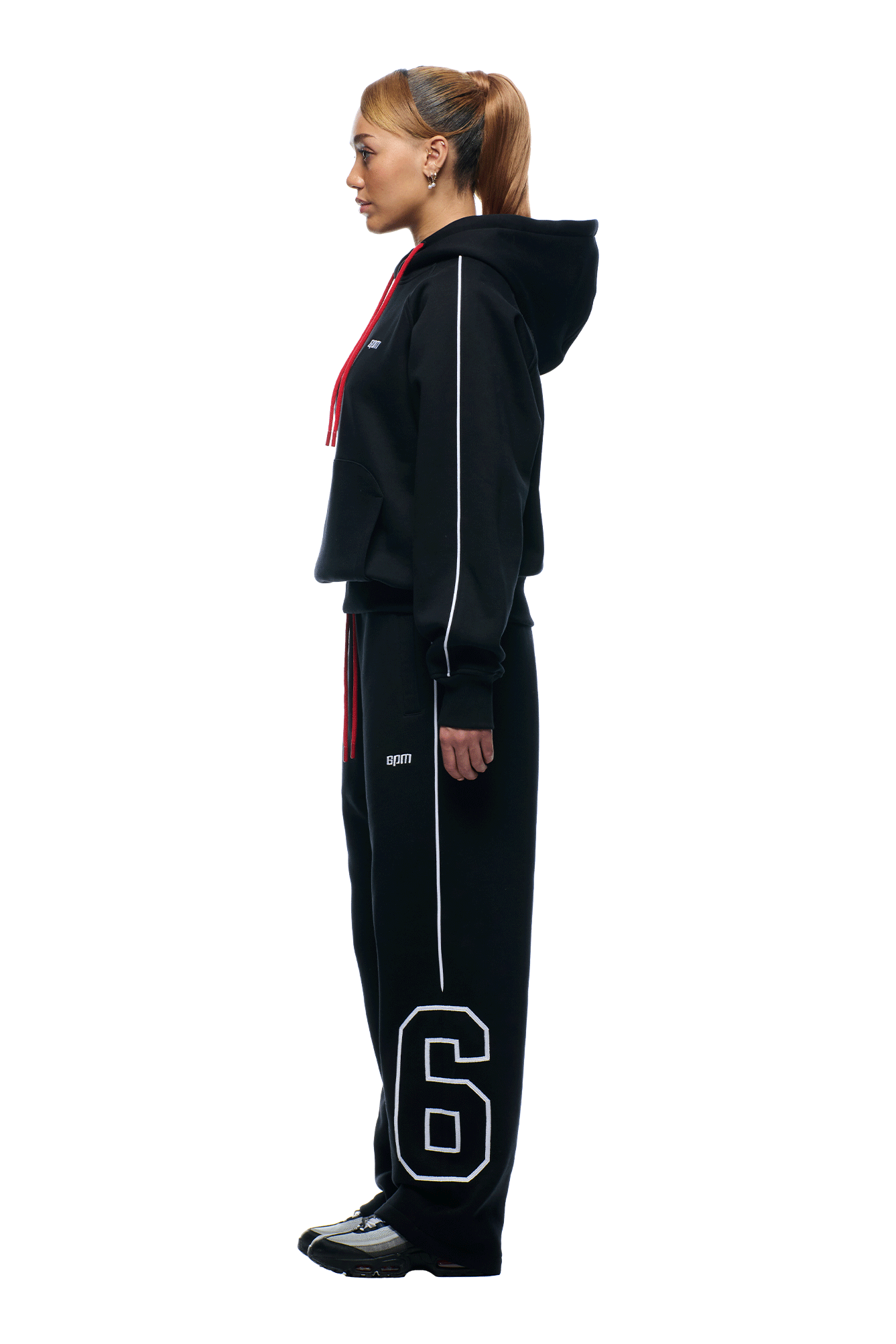 A woman in profile wears the NFL PIPING HOODIE and matching sweatpants from 6PM, featuring a number 6 on the left leg. She sports a high ponytail and black sneakers against a white background.