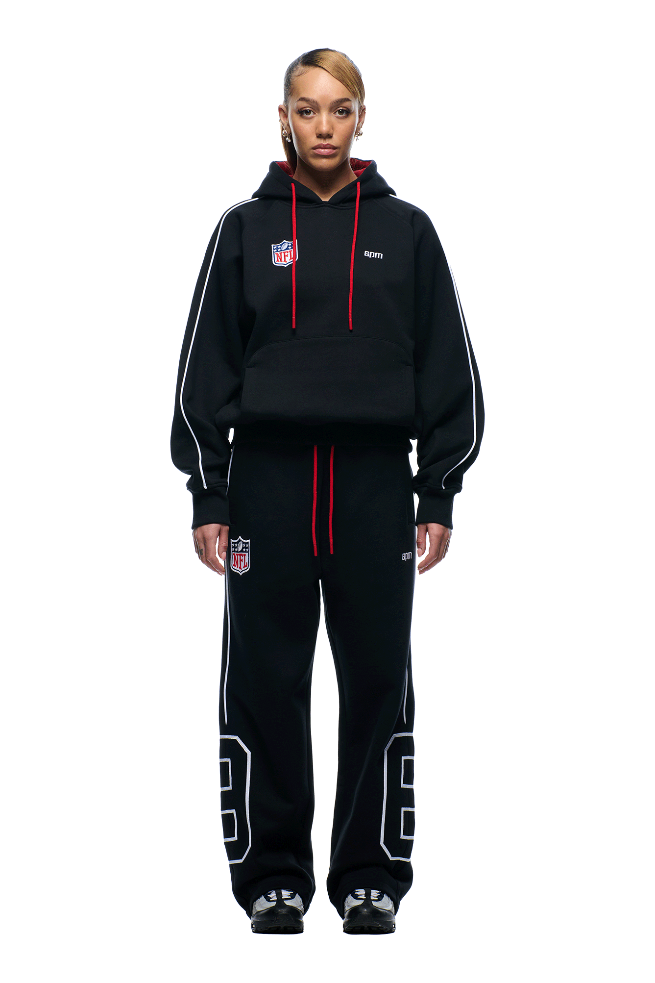 A person faces forward in a 6PM NFL PIPING HOODIE and matching black sweatpants with white stripes, red drawstrings, NFL logos, and large white numbers on the legs. The background is plain white.