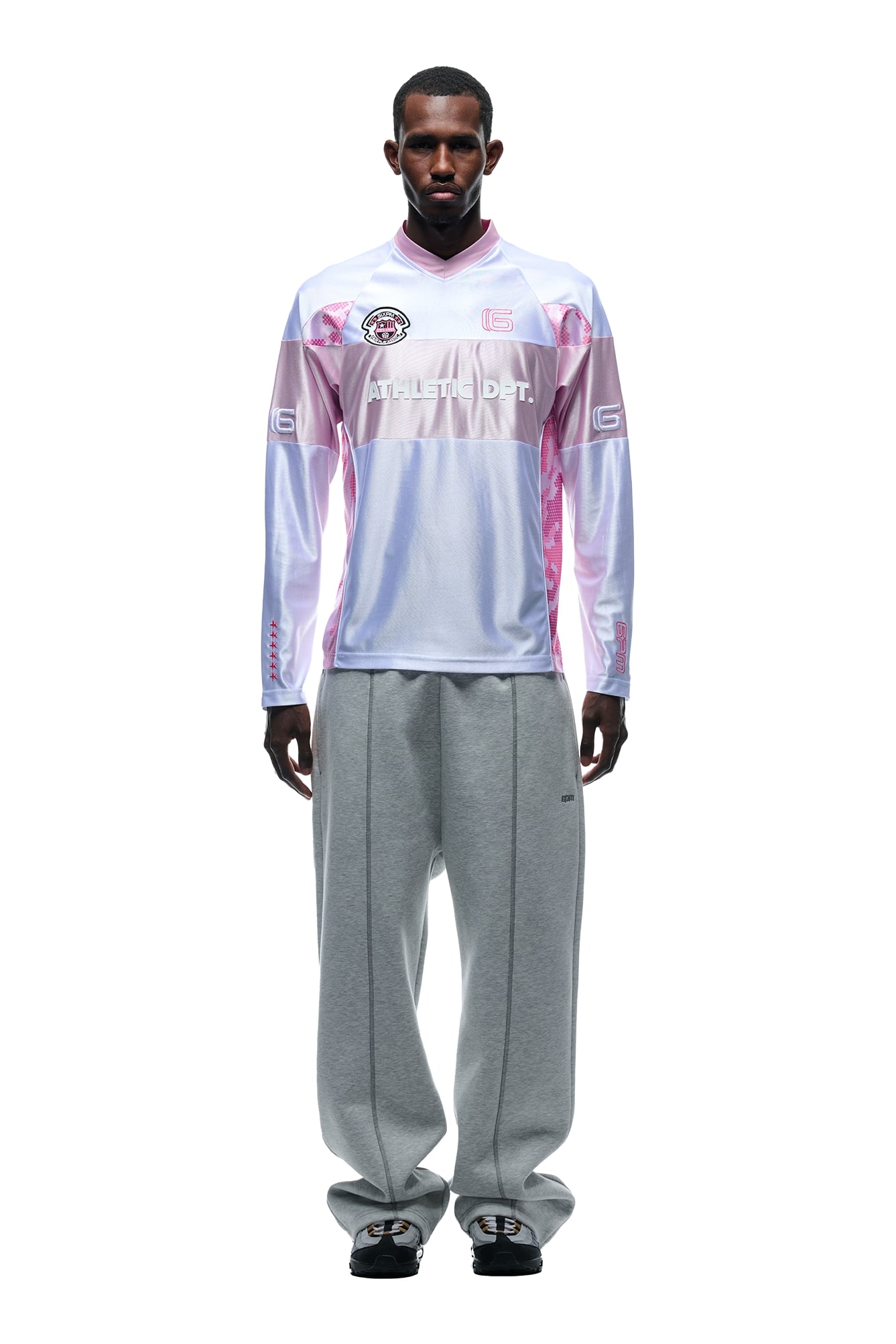 A man stands facing forward in a 6PM LONGSLEEVE JERSEY WHITE/PINK, paired with loose gray sweatpants and black shoes, against a white background.