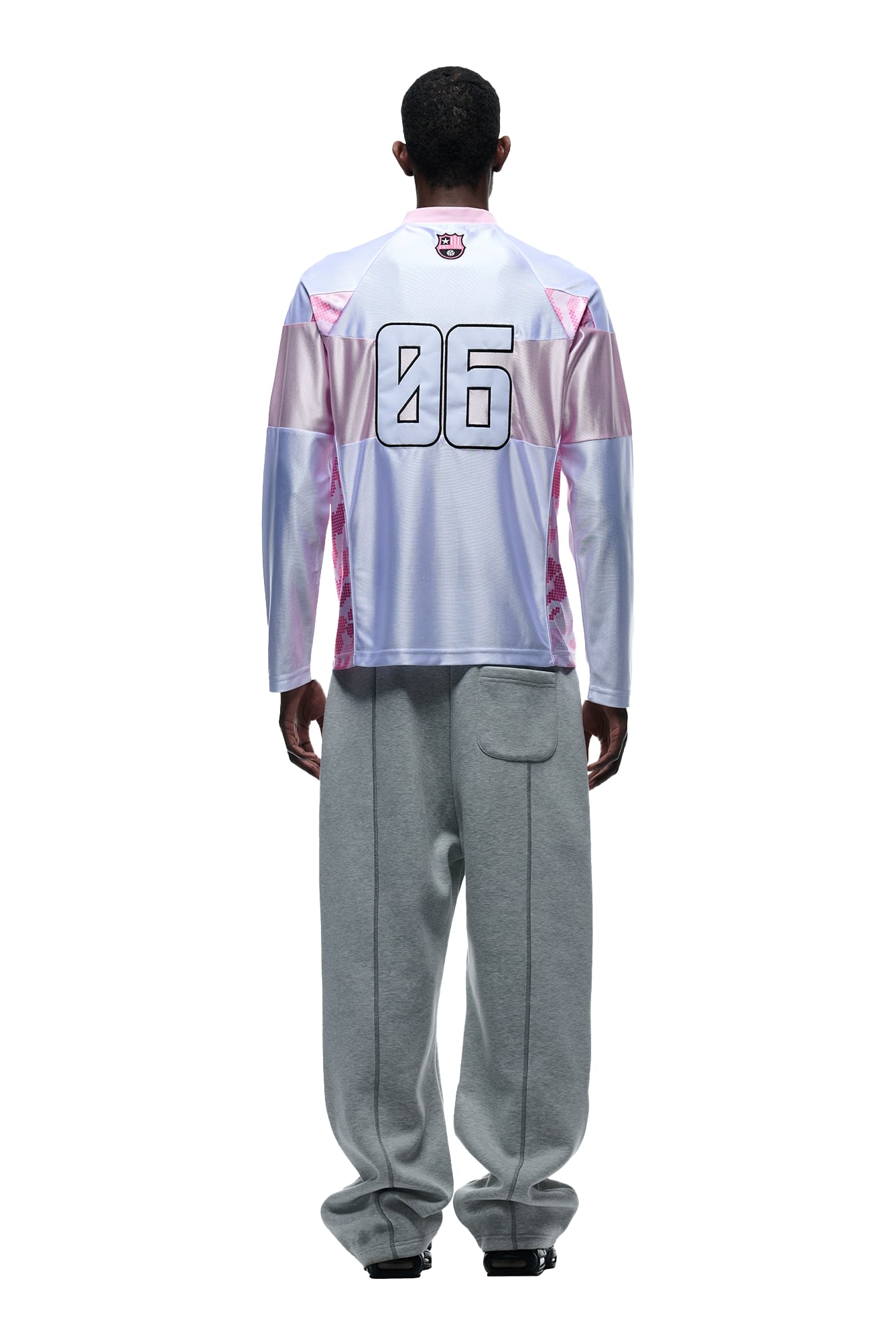 A person faces away, wearing a 6PM LONGSLEEVE JERSEY WHITE/PINK with the number 06 on the back, paired with loose gray sweatpants and black shoes, against a white background.