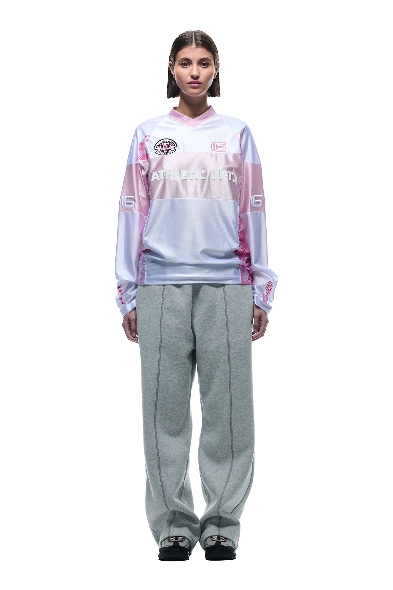 A woman faces forward in a 6PM LONGSLEEVE JERSEY WHITE/PINK with logos, light gray wide-leg sweatpants, and black shoes against a plain white background.
