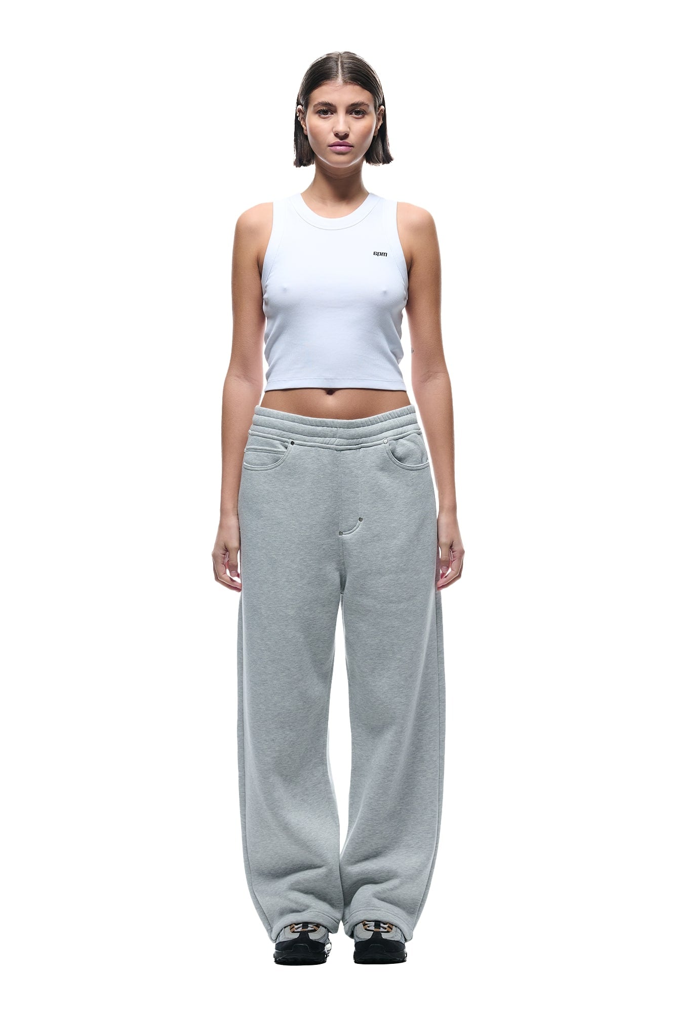 A woman with short brown hair wears 6PM’s X-TRA WIDE 5-POCKET JOGGER GREY sweatpants, a white sleeveless crop top, and black sneakers, standing against a plain white background with a neutral expression.