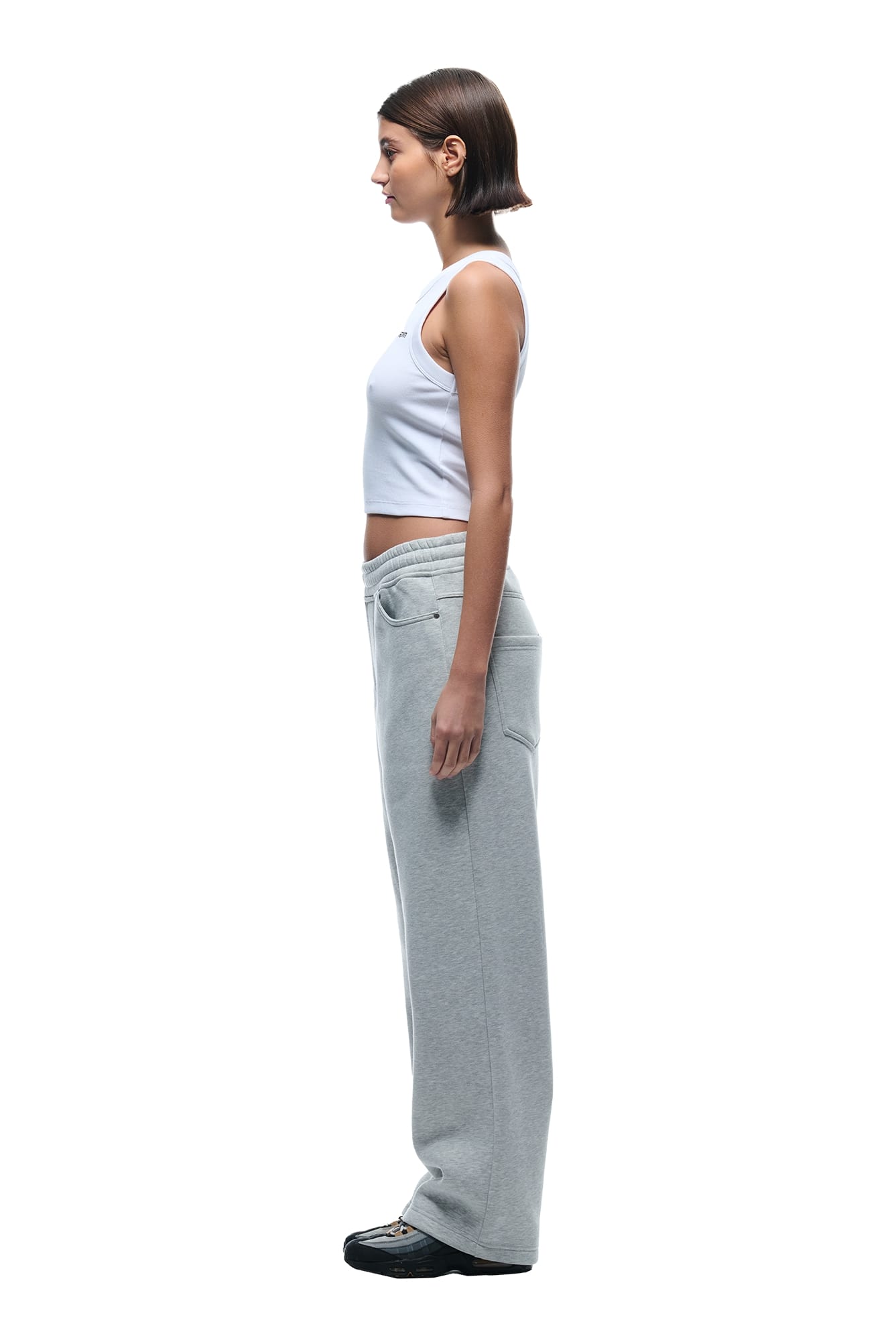A woman with short brown hair stands in profile against a plain white background, wearing the 6PM X-TRA WIDE 5-POCKET JOGGER GREY, a white sleeveless crop top, and black shoes.