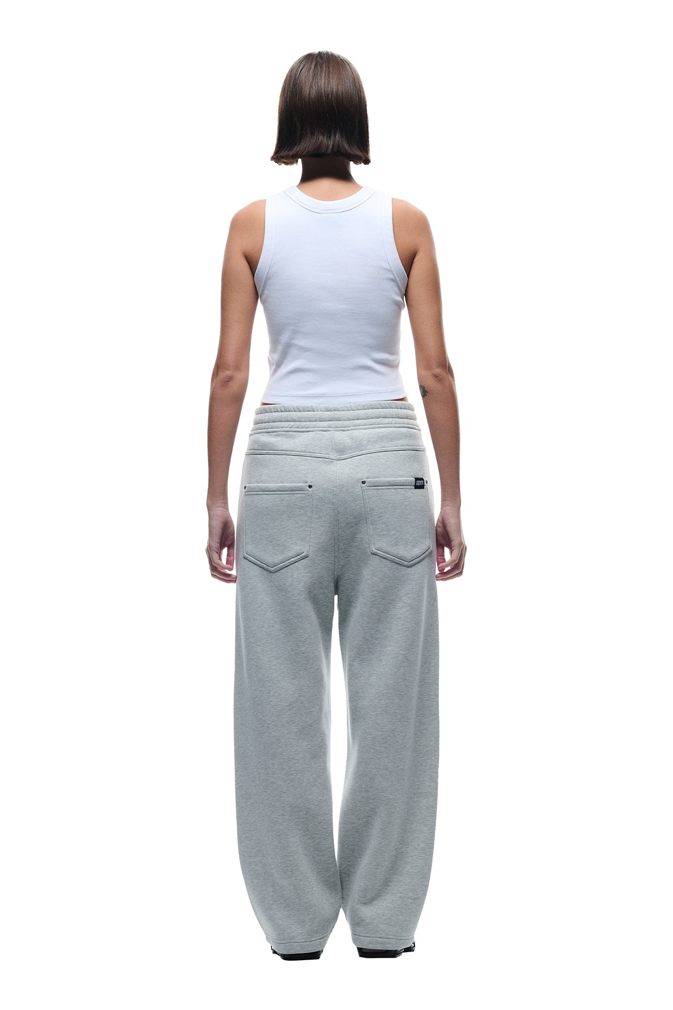 A person faces away from the camera, wearing a white sleeveless tank top and 6PM X-TRA WIDE 5-POCKET JOGGER GREY sweatpants. The background is plain white.